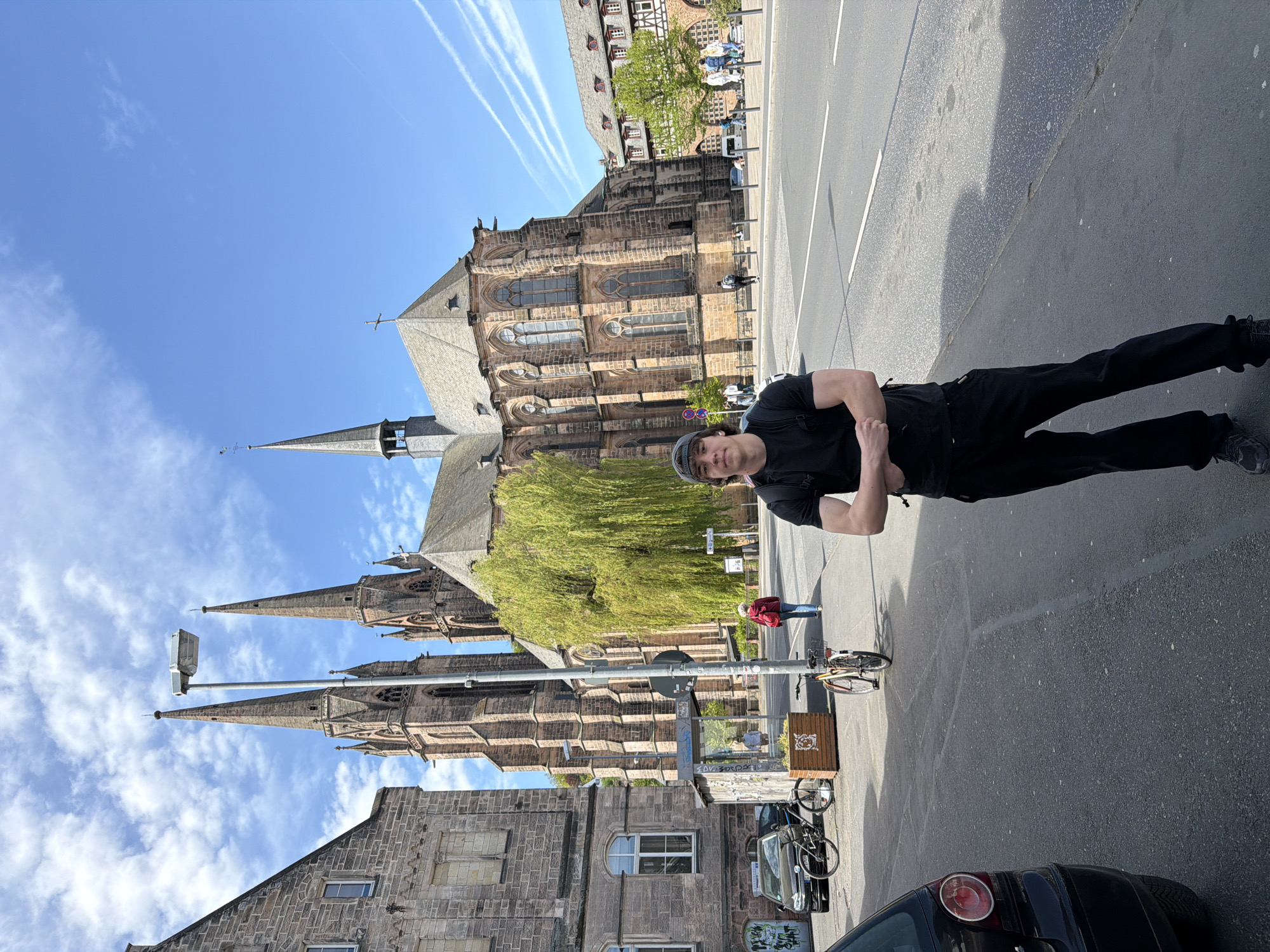 The image depicts a sunlit street scene in Marburg, Germany, featuring a prominent gothic church. In the foreground, a young man stands on the paved sidewalk, wearing a black t-shirt, dark pants, and a dark baseball cap. He has his arms crossed and looks towards the camera with a neutral expression. His shadow is cast on the ground to his right.
Behind the man, a wide street with white markings separates the foreground from the background. The dominant feature in the background is a large, ornate stone church with multiple spires, intricate architectural details, and arched windows, identifiable as St. Elisabeth Church. A vibrant green-leafed tree stands near the church. Other stone-built structures are visible to the left of the church and further down the street.
The sky is bright blue with several white contrails and scattered wispy clouds, indicating clear and sunny weather. The strong sunlight suggests a daytime setting, likely mid-morning or afternoon, given the length and direction of the shadows. Several other pedestrians are visible in the distance near the church and across the street, and bicycles are parked along the sidewalk and street. A black car is partially visible in the bottom right corner of the frame. A tall street lamp pole stands in the mid-ground. No discernible text is visible.