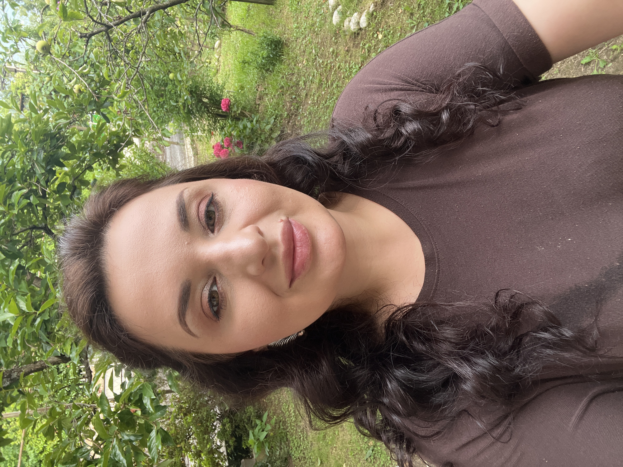 A medium shot shows a woman with long, dark, wavy hair and brown eyes, wearing a brown t-shirt. She is outdoors in a garden with green trees and grass, and pink flowers are visible in the background. The woman is looking at the camera and smiling. The location is Sokhumi, Abkhazia.