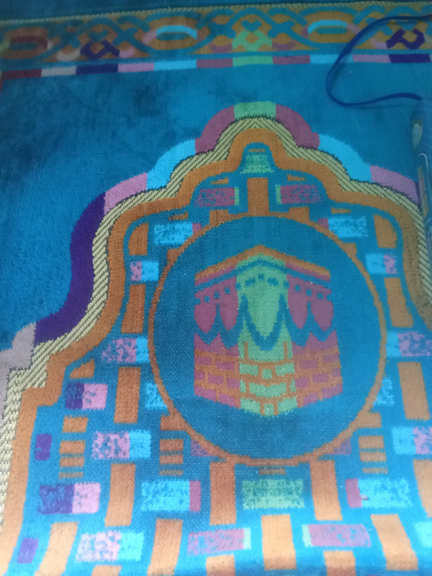 A close-up, high-angle shot shows a turquoise prayer rug with an intricate geometric pattern. The center of the rug features a circular motif depicting the Kaaba in Mecca, rendered in pink, orange, and green. The rug is adorned with various abstract shapes and patterns in shades of blue, purple, pink, orange, and yellow. A dark blue cord is visible snaking across the upper right portion of the rug. The image does not contain any information about the city of Cempaka, Indonesia.