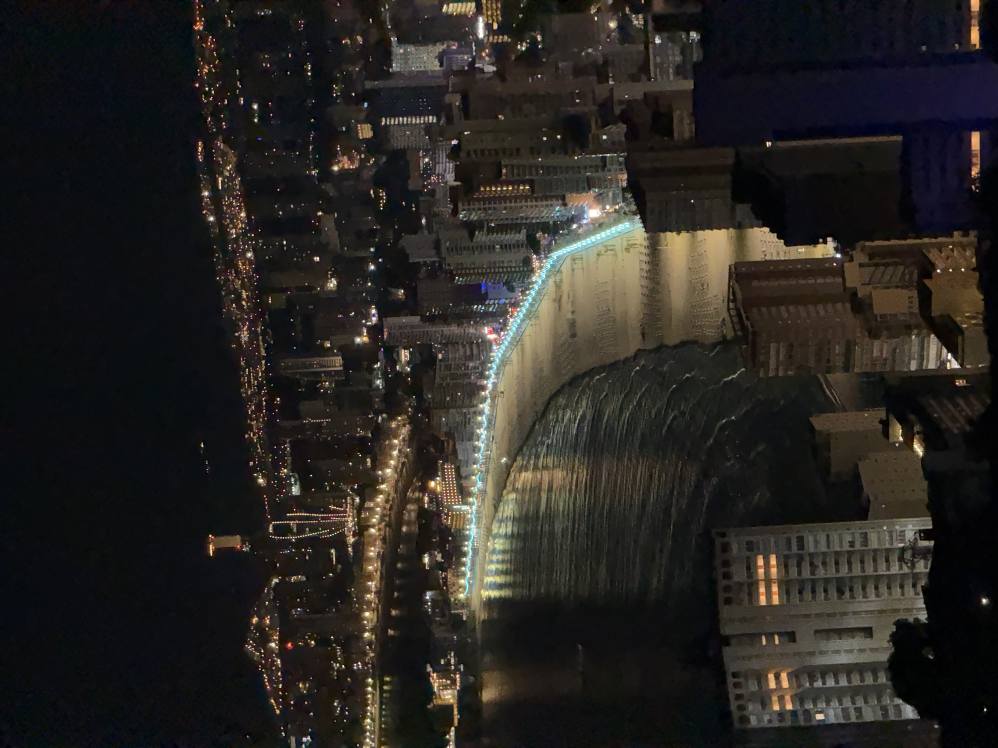 An aerial, nighttime view shows the Benidorm coastline in Spain. The beach, illuminated by lights from the promenade and surrounding buildings, curves along the shore. The Mediterranean Sea, with gentle waves, reflects the city lights. High-rise buildings, hotels, and apartments dominate the cityscape, with many windows lit. In the distance, the iconic Gran Hotel Bali stands tall, its illuminated spire visible. The image captures the vibrant atmosphere of Benidorm at night, with the beach and urban landscape illuminated by a myriad of lights.