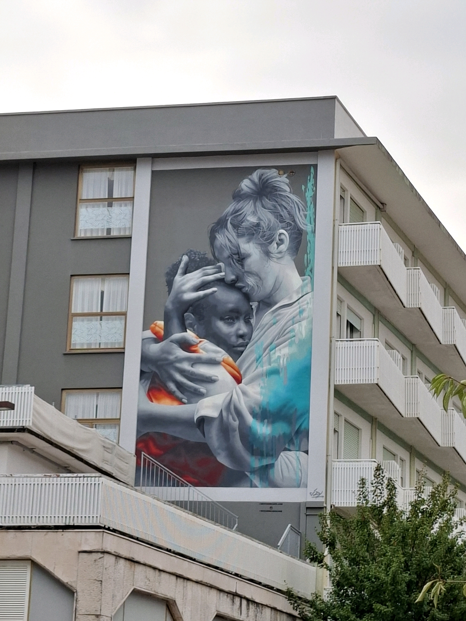 A mural depicting a woman embracing a child is painted on the side of a grey apartment building in Caorle, Italy. The woman, with her hair pulled up, cradles the child. The child, who has dark skin, is held close by the woman. Both figures are rendered in shades of grey and white, with splashes of turquoise and orange paint. The building has multiple balconies with white railings and windows covered by white curtains. Green trees are visible in the foreground, obscuring some of the lower levels of the building. The sky above is overcast.