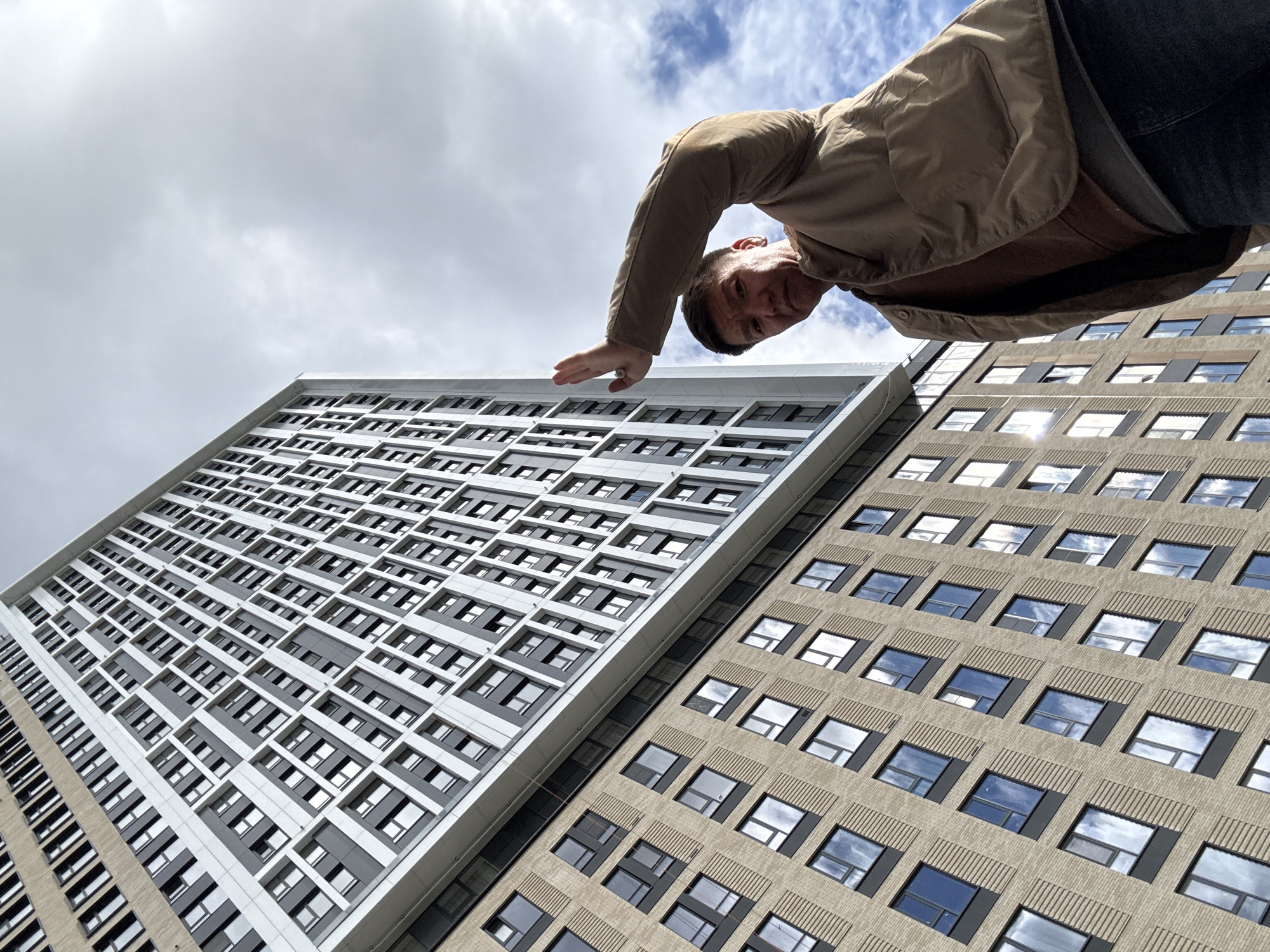 A low-angle shot shows a man leaning towards the camera with a backdrop of two adjacent buildings in Khimki, Russia, under a partly cloudy sky. The man is wearing a tan jacket over a brown shirt and dark jeans. He is positioned at the top right of the frame, with his head tilted down and his left hand extended towards the left side of the image. The buildings are tall and rectangular, filling most of the frame. The building on the left has a grid-like facade with alternating gray and white rectangular panels. The building on the right has a more traditional design with rows of square windows. The sky is visible in the upper left corner, with white clouds against a blue background.