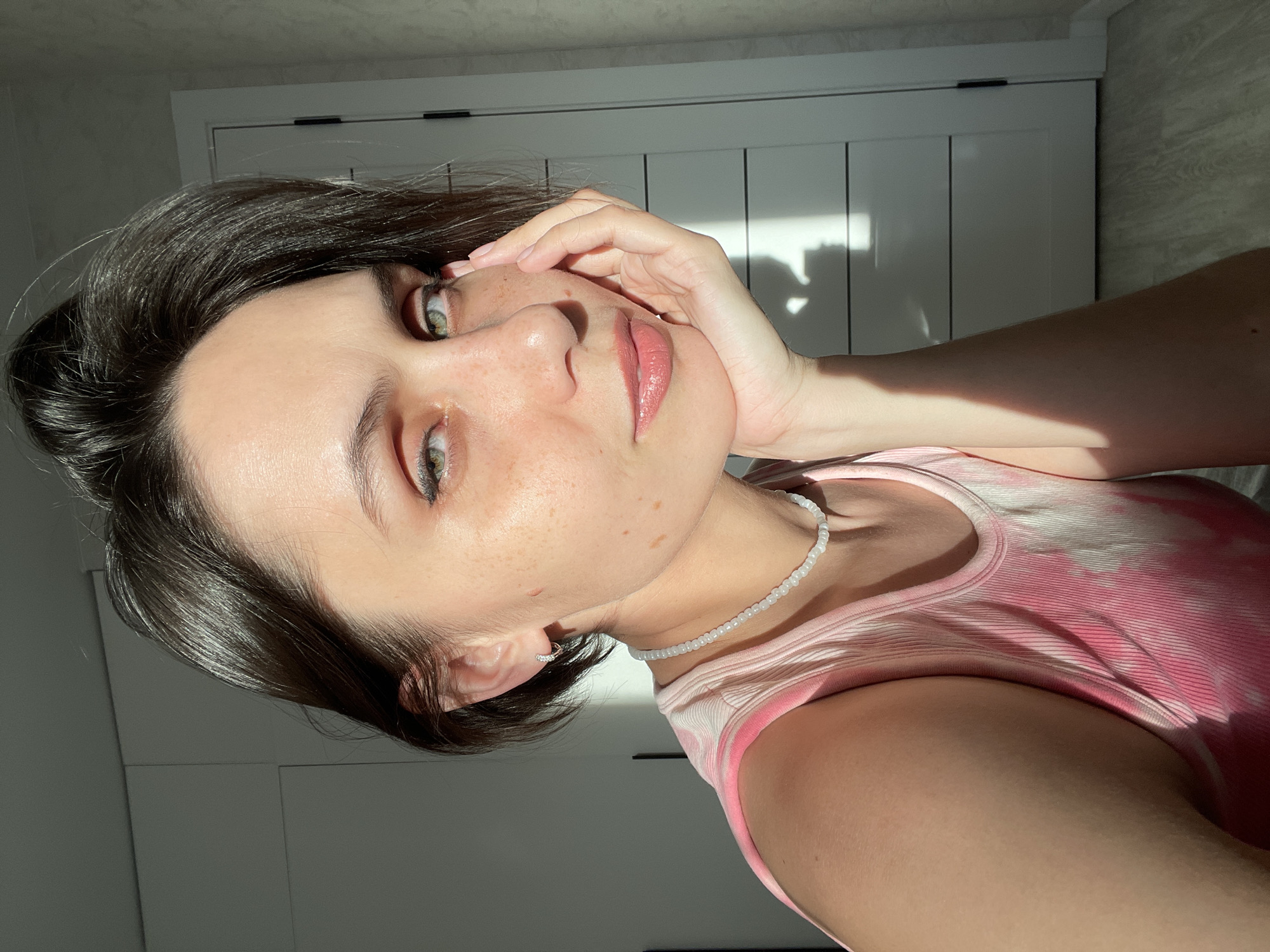 A young woman is facing the camera, her head resting on her left hand. She has short, dark brown hair and fair skin with freckles. She is wearing a pink tie-dye tank top and a white beaded necklace. The background consists of a white door with vertical panels and a partially visible white wall. Sunlight is cast upon the scene, creating shadows on the door. The location of the image is not identified and is therefore not in the city of Izhevsk, Russia.