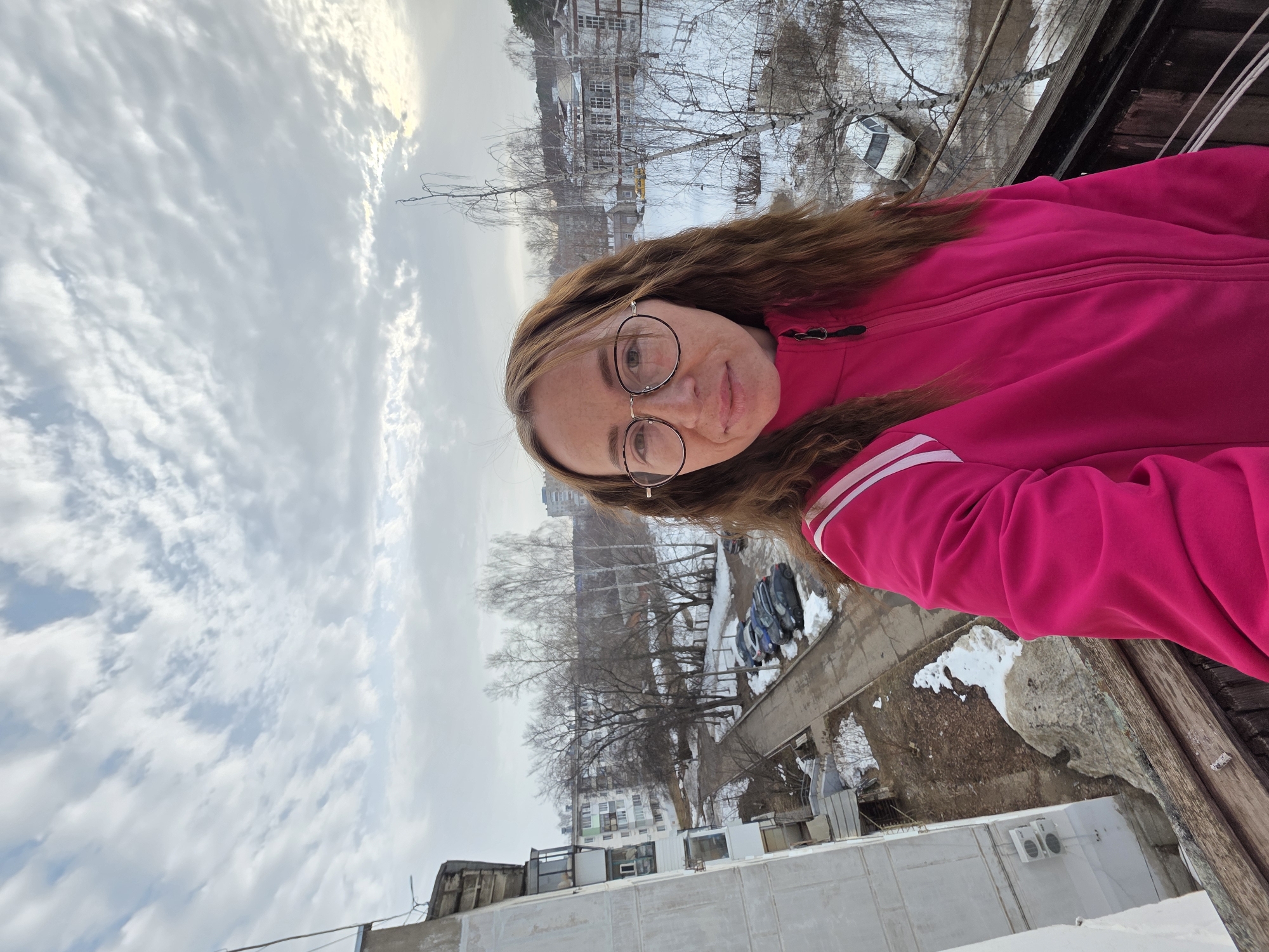 A female subject, appearing to be in her mid-30s to early 40s, with light brown, wavy hair and round, gold-rimmed eyeglasses, is depicted in a selfie, looking directly at the camera with a slight, neutral smile. She is wearing a bright pink zipped athletic jacket with white stripes on the shoulders. The subject is positioned outdoors from an elevated perspective, likely a balcony or upper floor, with a weathered wooden railing in the immediate foreground.
The background reveals an urban environment in Chaikovsky, Russia, characterized by late winter or early spring conditions. To the left, a multi-story residential building with balconies and two external air conditioning units is visible. A paved road or pathway traverses the mid-ground, bordered by patches of snow and exposed brown earth. Several vehicles, including dark sedans and a white car, are parked along this road. Numerous bare deciduous trees are dispersed throughout the scene, interspersed with additional multi-story residential buildings and a complex of older, multi-windowed structures in the distance to the right. The sky is overcast with textured white and grey clouds.