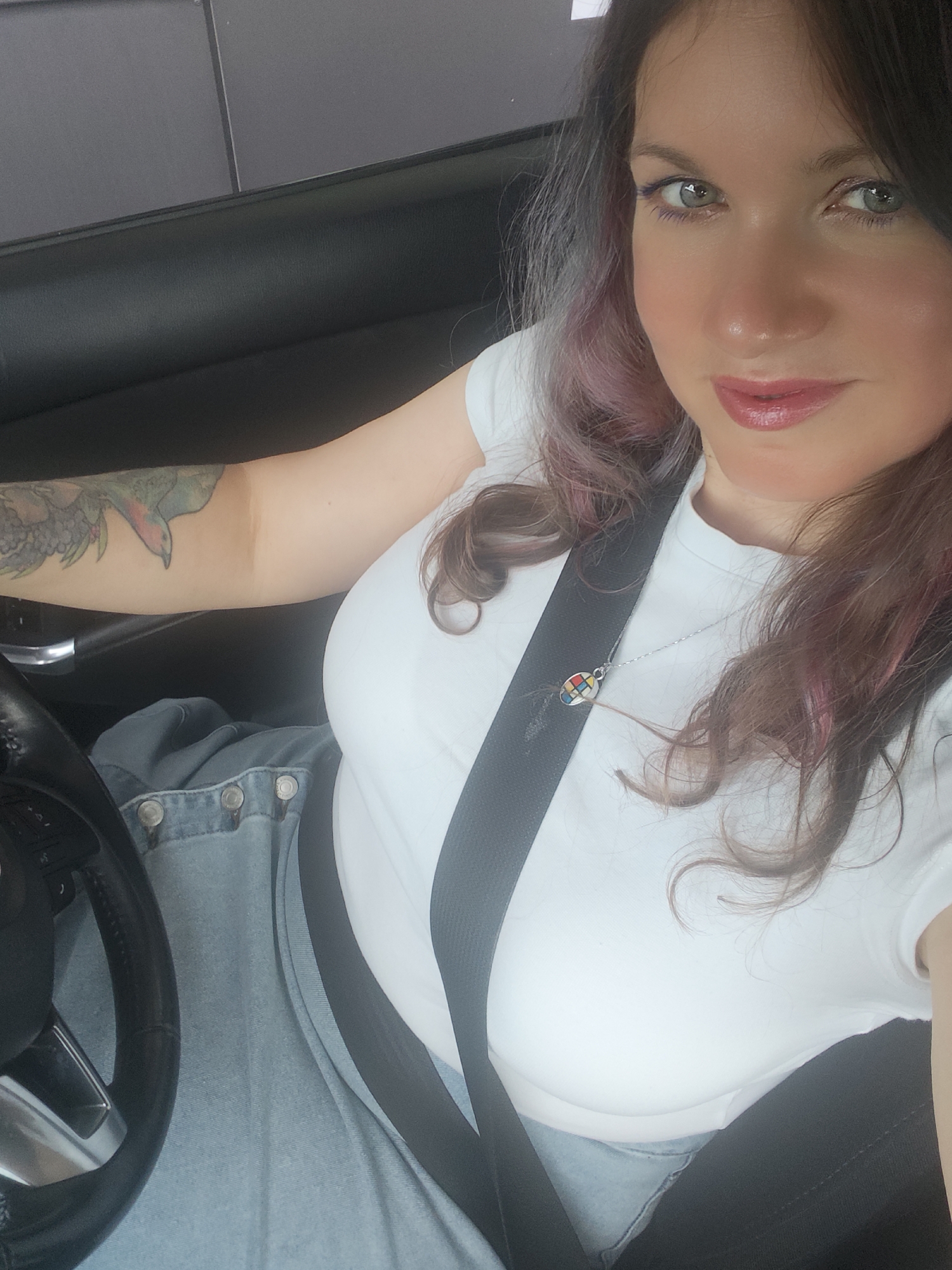 A woman is seated in the driver's seat of a car, with her right arm on the steering wheel and her left arm resting on her lap. She is wearing a white t-shirt and a denim jacket. A seatbelt is fastened across her chest. She has long, dark hair with some pink streaks. She is looking at the camera and smiling. A tattoo of a bird is visible on her left arm. She is wearing a necklace with a colorful, abstract pendant. The interior of the car is dark, with the steering wheel and dashboard visible. The image appears to have been taken during the daytime, possibly in a shaded area, given the lighting. The overall mood is casual and relaxed.