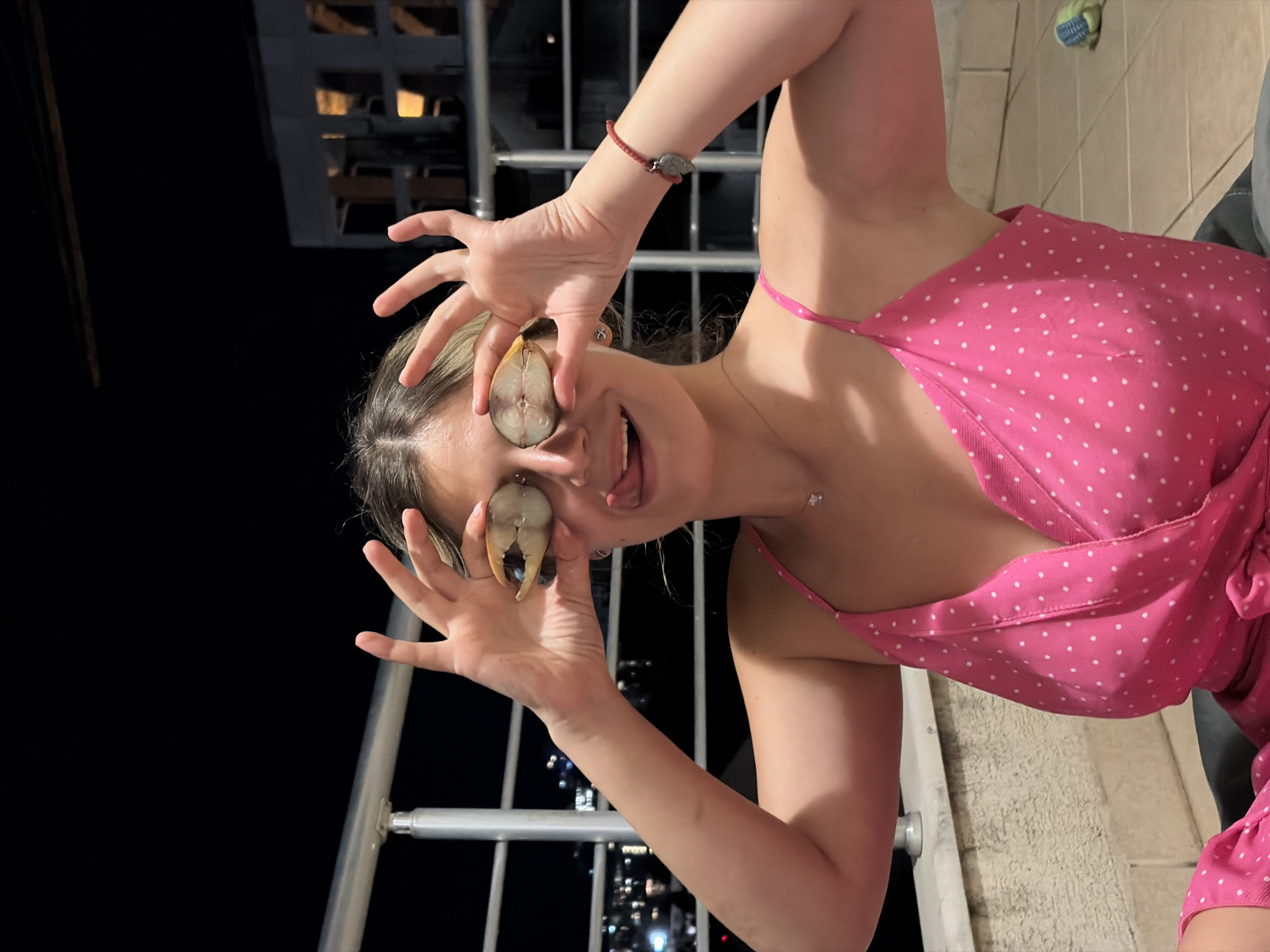 In the image, a young woman with blonde hair is sitting on a balcony at night in Budva, Montenegro. She is wearing a pink polka dot dress and holding two slices of fruit over her eyes, sticking her tongue out playfully. The background shows a dimly lit cityscape with buildings and a railing.