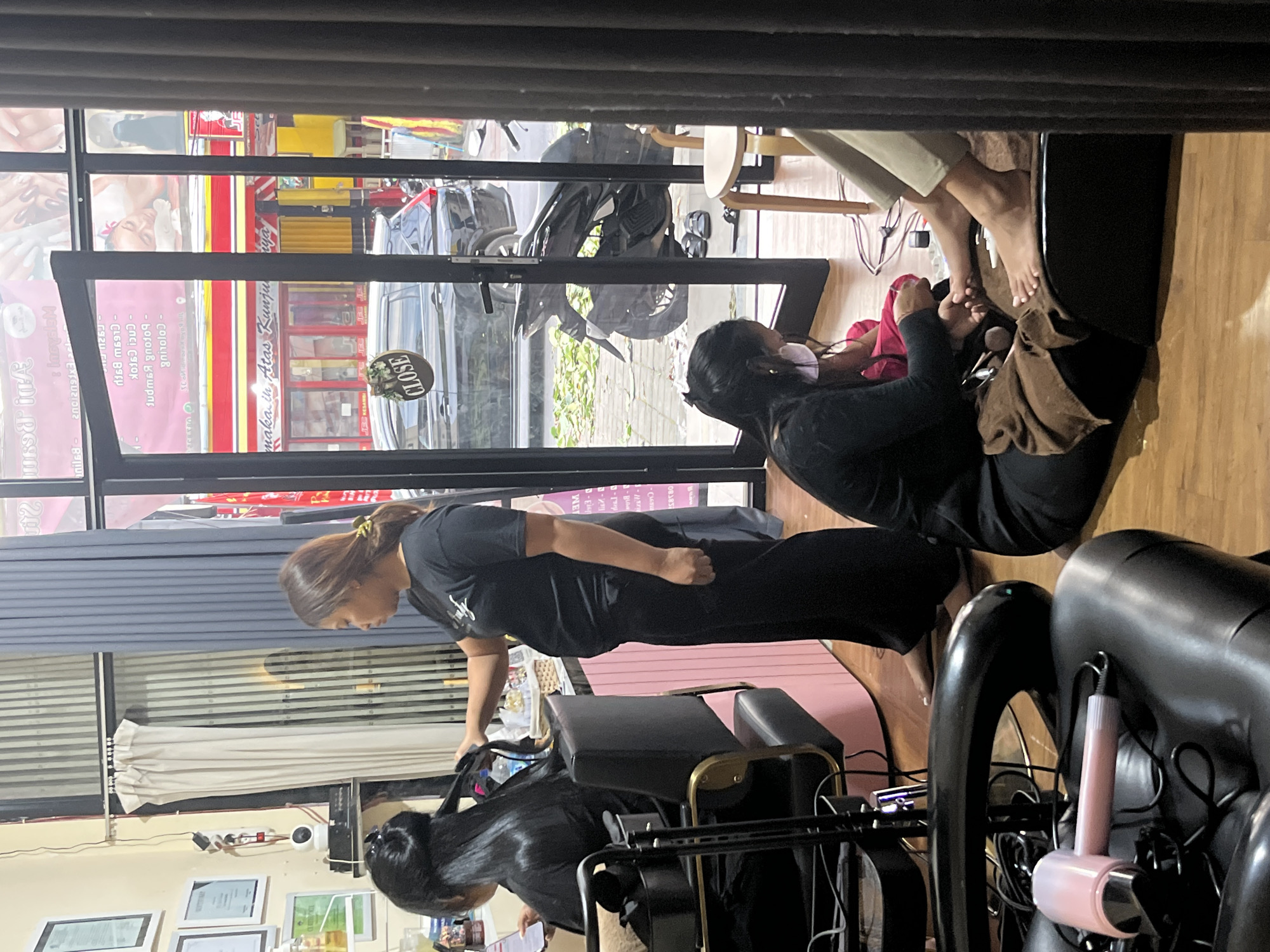 This media file shows the interior of a salon or spa. Two women are visible. One woman, with dark hair and wearing a black top and pants, is seated and appears to be receiving a foot massage from another woman who is kneeling. The kneeling woman has dark hair and is wearing a black outfit. Her hands are focused on the seated woman's feet.
In the background, through a glass window, a street scene is visible. There are several motorcycles and a car parked outside, suggesting it's daytime. Signs with text, some in Indonesian, are visible on the storefronts. One sign in the window clearly reads "CLOSE". The salon interior appears to be clean and organized, with chairs and equipment suggesting services like hair styling or nail care. The overall atmosphere seems relaxed, consistent with a spa or salon setting. There is a pink hair dryer tool visible in the foreground.