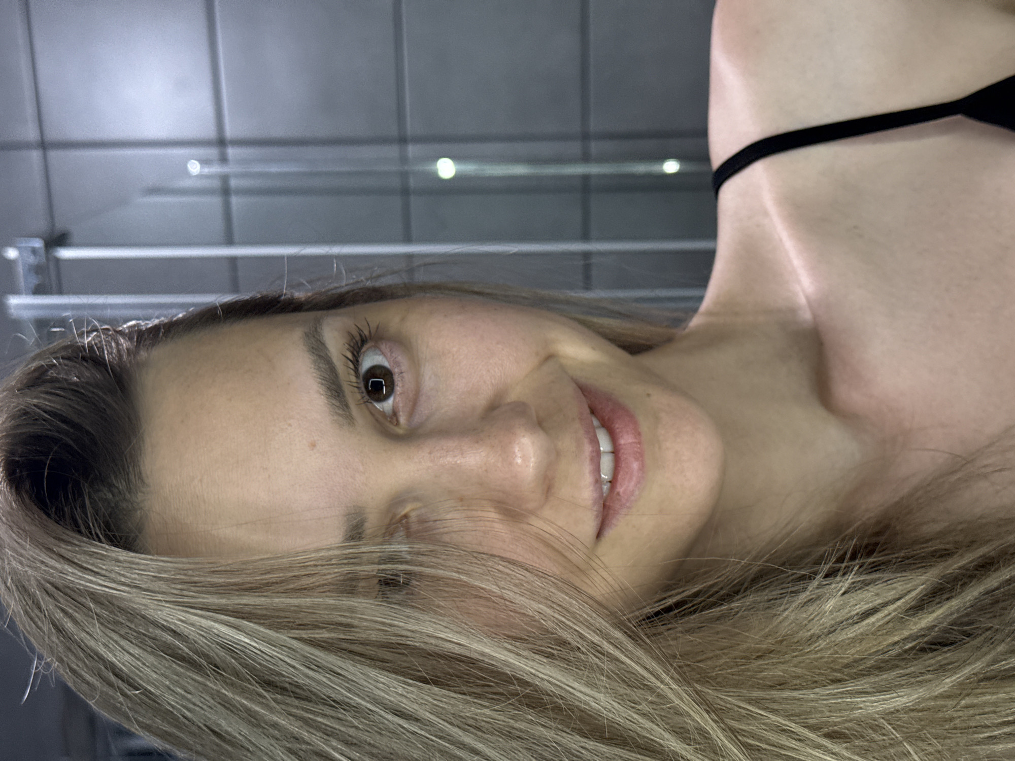 A medium close-up shot depicts a woman with light blonde highlighted hair in an interior setting. She is positioned at an angle, with her face tilted slightly towards the camera. Her eyes are brown and she has a gentle smile, revealing her teeth. Part of a black strap is visible, suggesting clothing. The background consists of tiled walls, possibly a bathroom. The location is unknown.