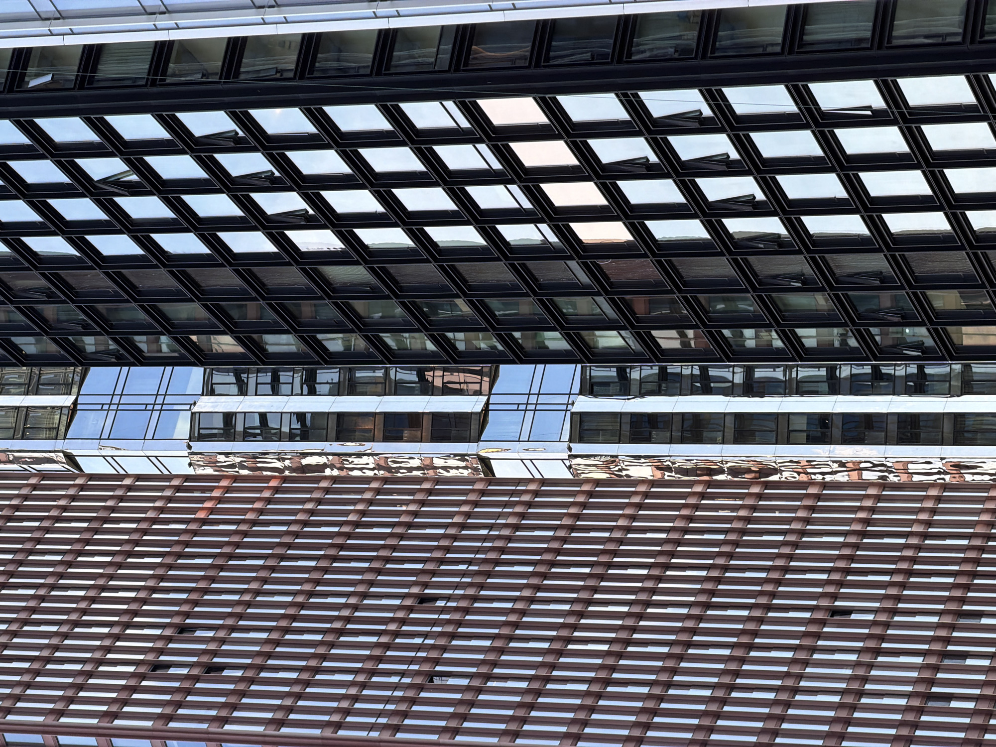 The image shows a detailed view of architectural elements in Moscow, Russia. The scene features multiple layers of building structures. At the top, there is a section with a grid-like pattern of windows, some of which are slightly ajar, reflecting the sky and surroundings. Below that is another layer of glass and metal architecture, reflecting an adjacent building with rows of windows. In the foreground, a brown horizontal structure with repeating lines partially obscures the layers behind it. The structures are modern and appear to be located in an urban environment.
