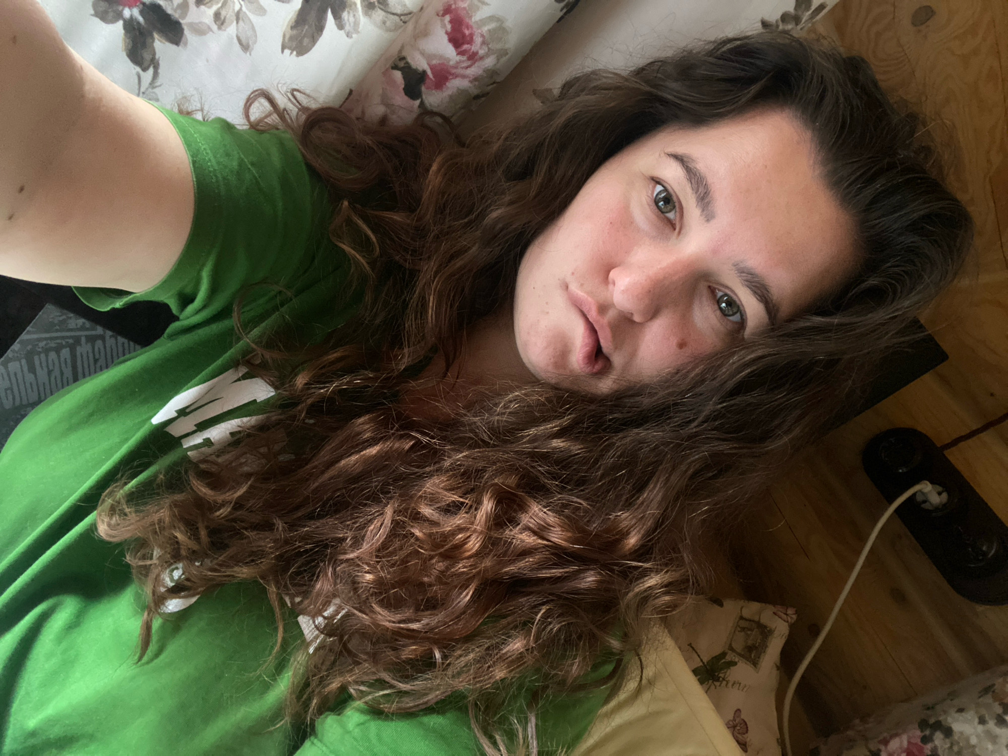 The image shows a young woman lying on her side, looking directly at the camera. She has long, wavy brown hair that falls around her face and shoulders. She is wearing a green t-shirt. Her left arm is bent at the elbow and partially visible. The background appears to be a bed with floral patterned pillows and a wooden surface. A black electrical device with a white cord is visible in the lower right corner. The scene is indoors, likely a bedroom. There are no identifiable landmarks or specific details that would allow for a precise location within the city of Perm, Russia.