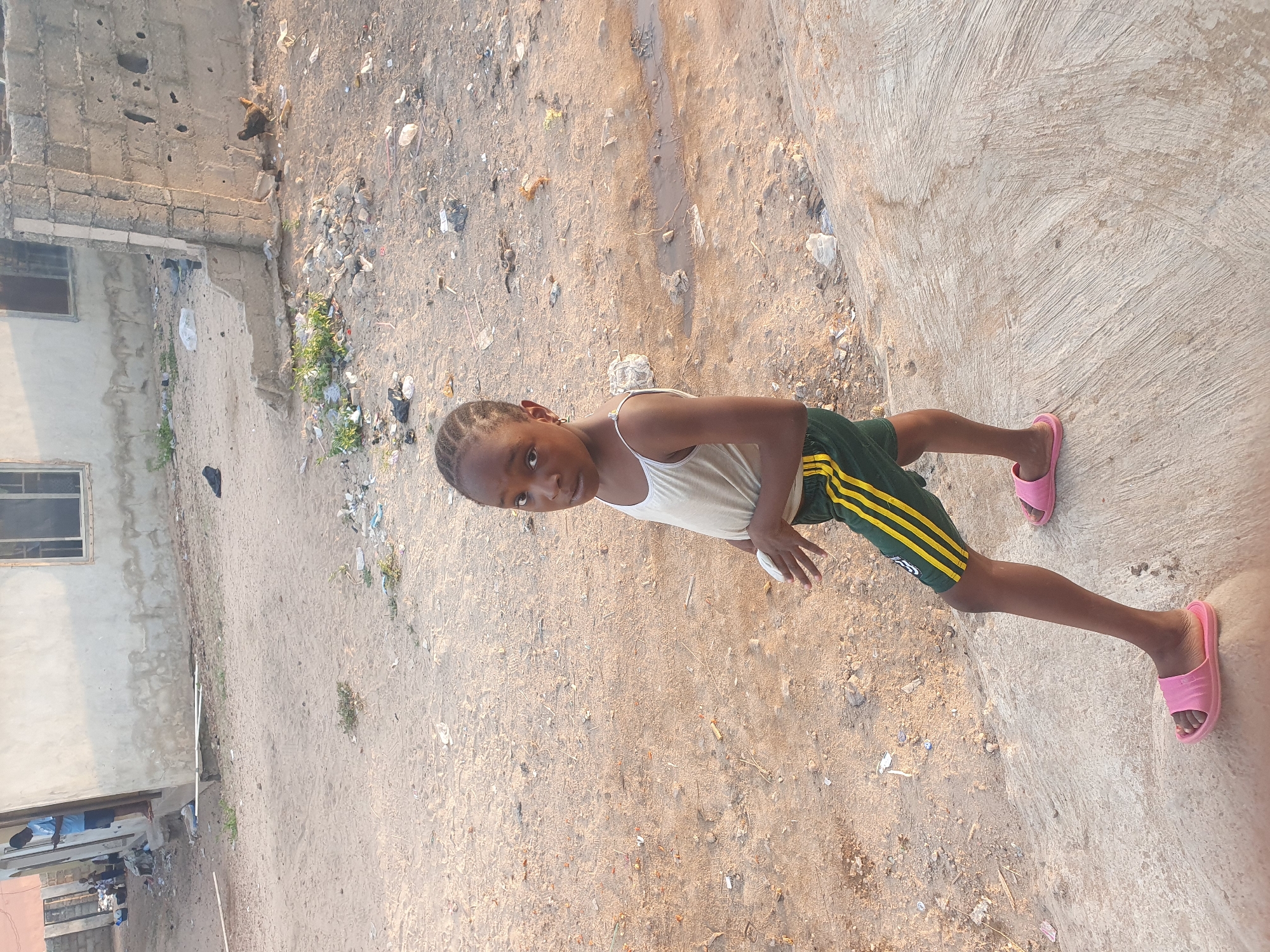 A young child stands in the center of the frame, looking directly at the camera. The child, likely a girl, wears a white sleeveless top, green shorts with yellow stripes along the sides, and pink slide sandals. She has braided hair.
The setting appears to be an outdoor, somewhat unpaved area. The ground beneath the child is a rough concrete slab, while the area behind her and to her left transitions into a sandy or dirt path. This path is littered with various debris, including plastic bags, small pieces of trash, and some rocks. Small patches of green vegetation sprout from the ground.
In the background, to the left, there is a building with unpainted concrete block walls and several windows. Part of another building or structure is visible further back. A chicken can be seen foraging near the wall in the mid-ground. The lighting suggests it is either late afternoon or early morning, with warm, soft sunlight illuminating the scene. The overall atmosphere is casual, and the child's expression is neutral and observant. There is no visible text in the image. The scene is consistent with an outdoor environment in a residential or semi-urban area, as suggested by the Lokoja, Nigeria context.