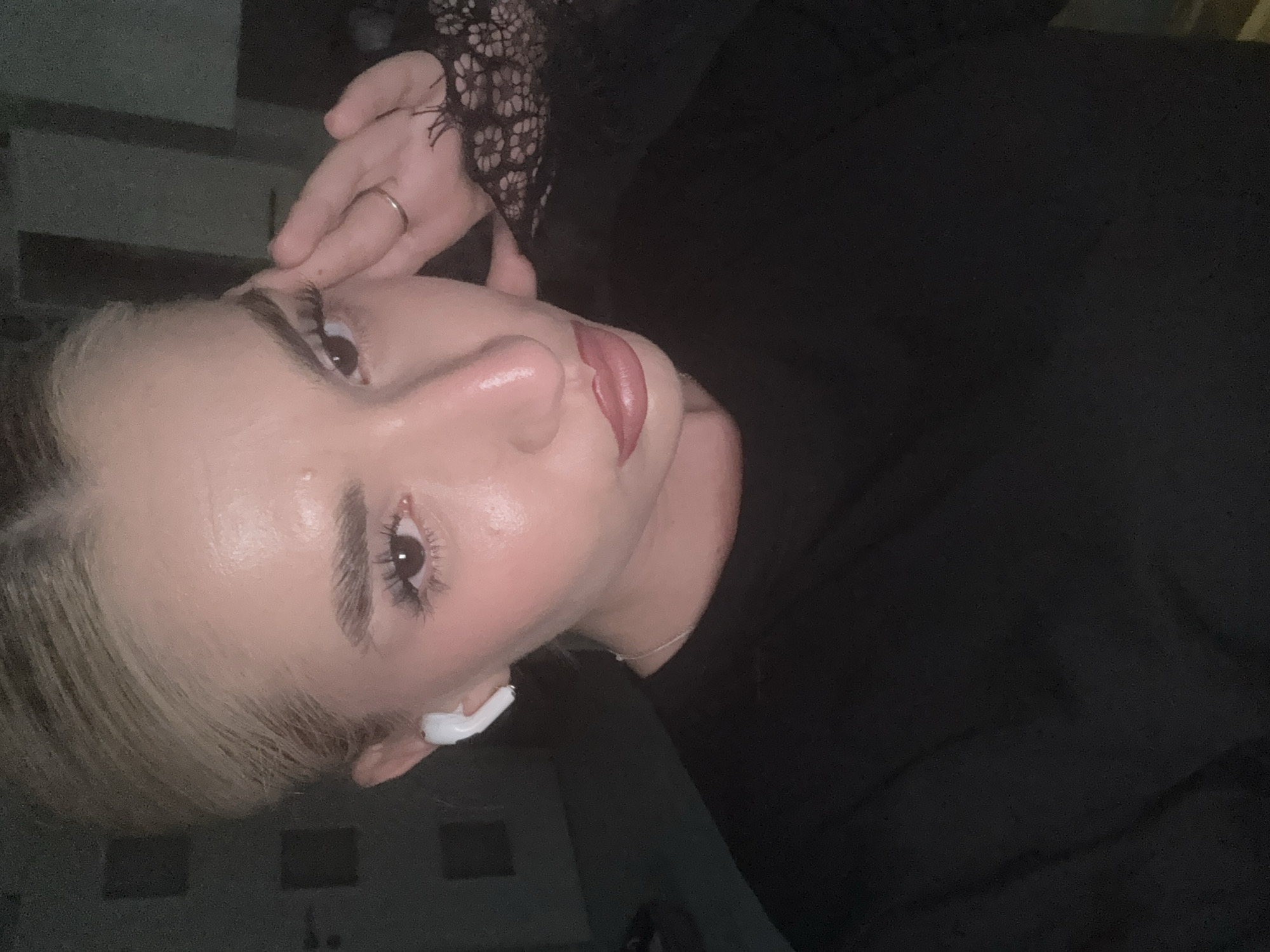 A close-up, tilted portrait captures a young woman from the chest up in what appears to be an indoor, low-light setting. She has blonde hair, neatly styled eyebrows, and prominent eyelashes. Her lips are a muted pink, suggesting lipstick, and her skin has a dewy finish. A white earbud (AirPod) is visible in her right ear. Her right hand, adorned with a simple silver-colored ring on her ring finger, is gently placed near her temple, with hints of a black lace sleeve or top. She is wearing a dark, possibly black, garment. The background is uniformly dark, offering no specific details about the environment. The image gives the impression of a posed selfie or portrait taken in the evening or at night. There is no visible text. While the location context is Sofia, Bulgaria, this is not discernible from the image itself.