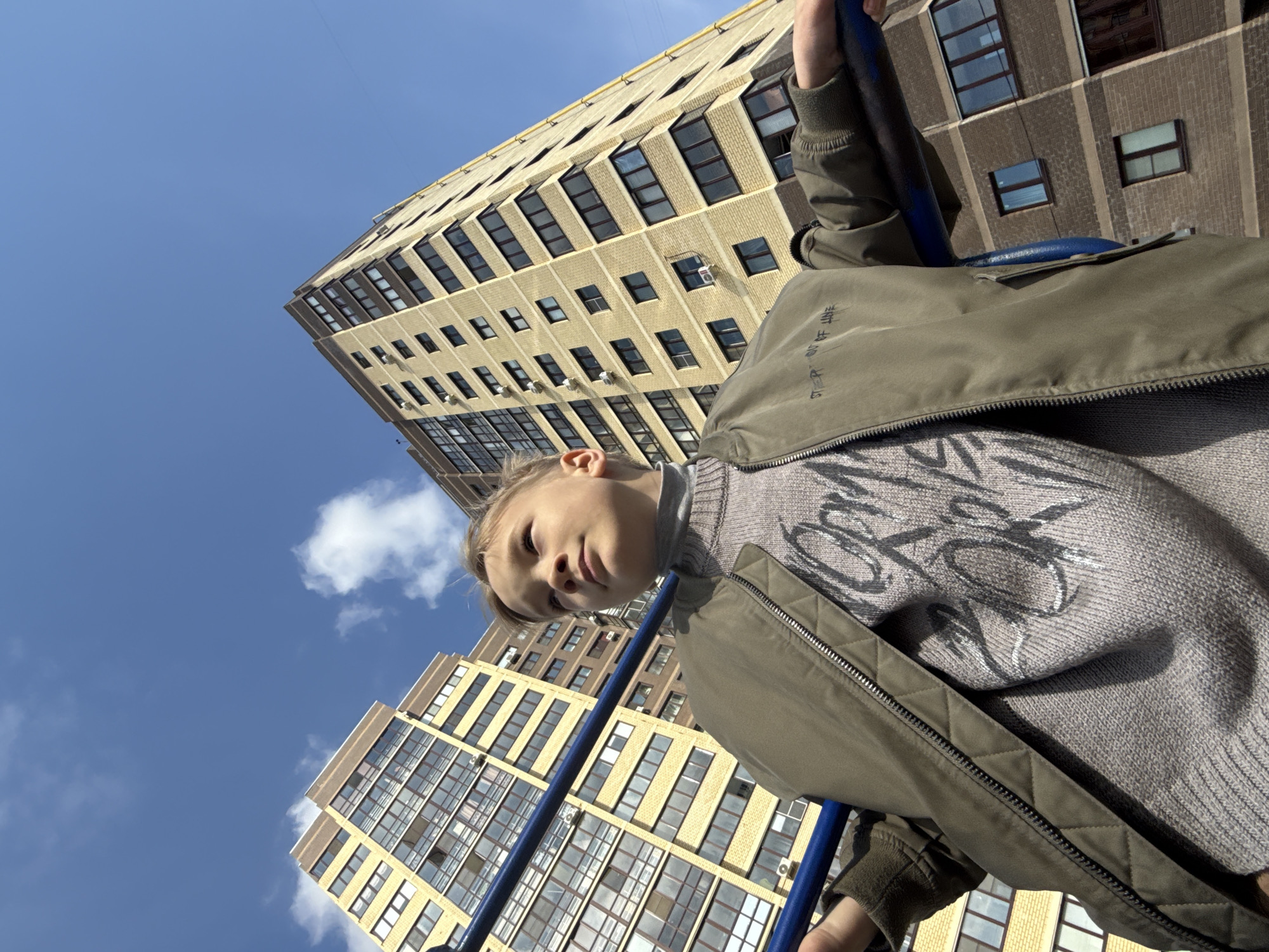 A low-angle shot shows a person in the lower foreground, wearing a gray sweater with a design, and a green jacket. Two high-rise buildings frame the shot in the background under a partly cloudy, bright blue sky. The buildings appear to be residential apartments, with numerous windows and a light-colored facade. The image was taken in Dmitrov, Russia.