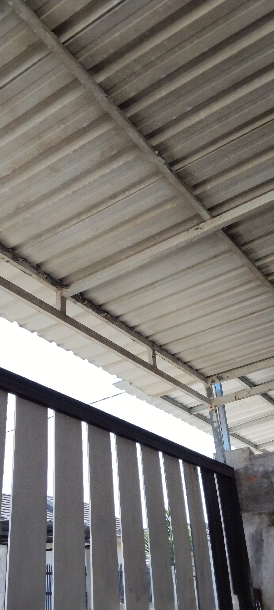 The image shows a corrugated metal roof with a supporting metal frame, likely part of a carport or outdoor structure. The roof is slightly weathered. Below the roof, there is a fence made of vertical concrete slabs framed by a dark metal bar. Through the gaps in the fence, parts of a building with a similar corrugated roof and some vegetation can be seen. The scene appears to be outdoors, possibly in a residential area, given the context of a fence and a building. The lighting suggests it is daytime. The location is identified as Duman, Indonesia. There are no people visible in the image.