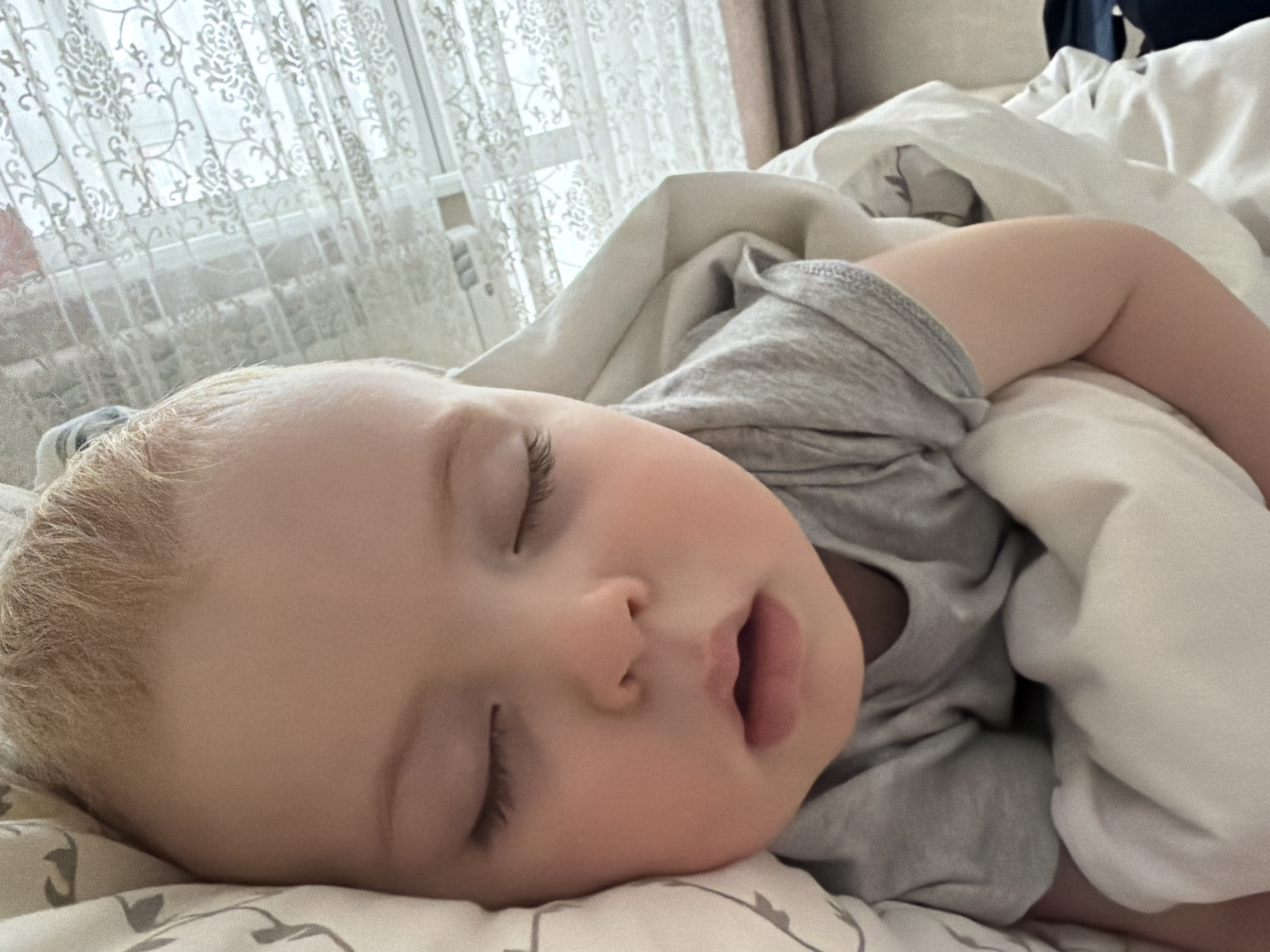 A young child with blonde hair is sleeping soundly in a bed covered with white bedding and a patterned pillowcase. The child is wearing a gray shirt. A patterned white curtain hangs near a window, and a partially visible curtain is in the background. This scene appears to be taking place indoors, likely in a residence. The precise location in Votkinsk, Russia, cannot be determined from the image alone.