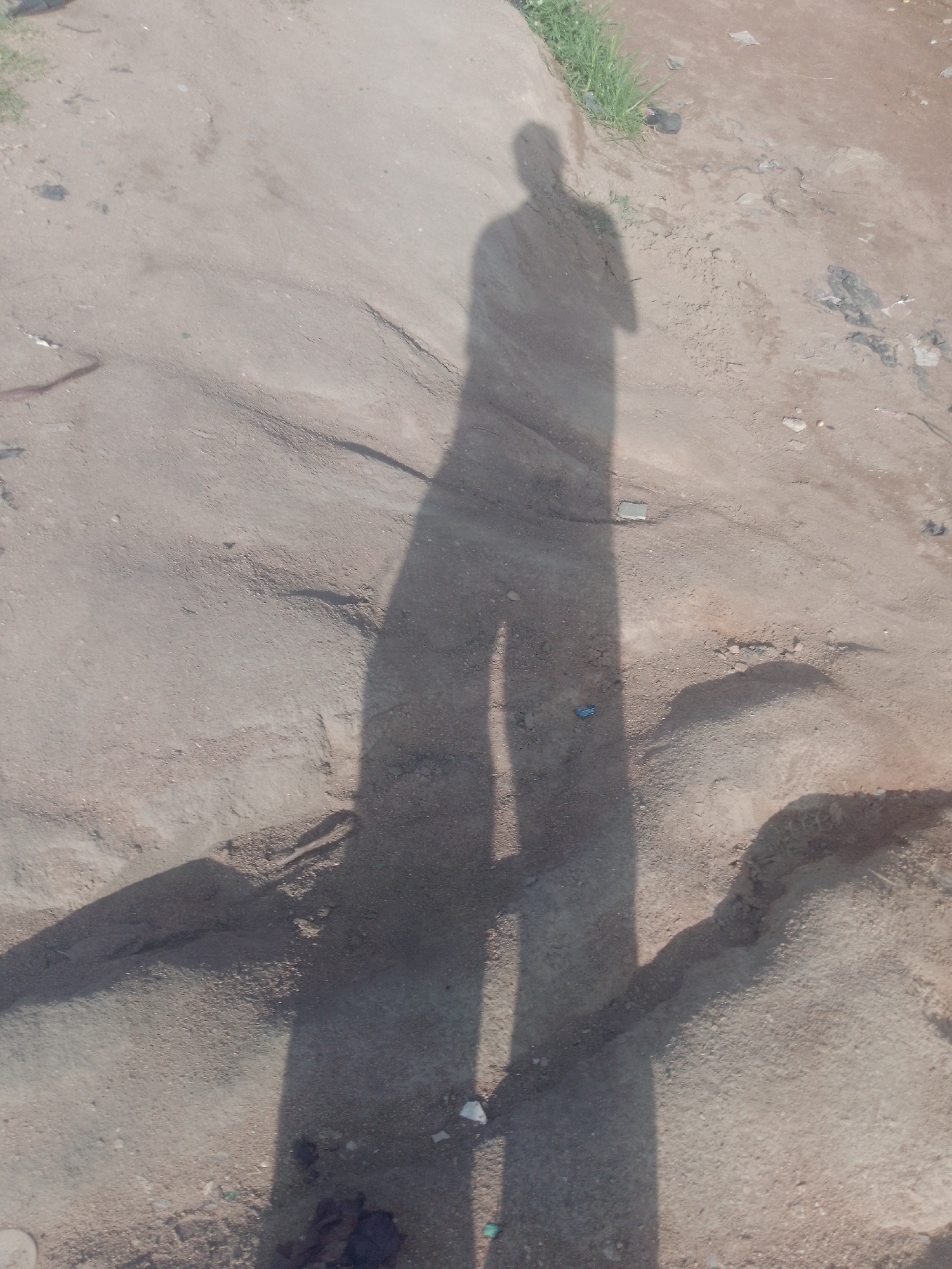 In Jalingo, Nigeria, a long shadow of a person is cast across a dusty, uneven ground. The shadow stretches from the top left of the image towards the bottom right. The ground is a mixture of sand and small pebbles, with some larger rocks and debris scattered about. In the top left corner, a patch of green grass can be seen, indicating the presence of vegetation. The overall impression is one of an outdoor, possibly unpaved area, bathed in sunlight, with the photographer's shadow being the primary subject.