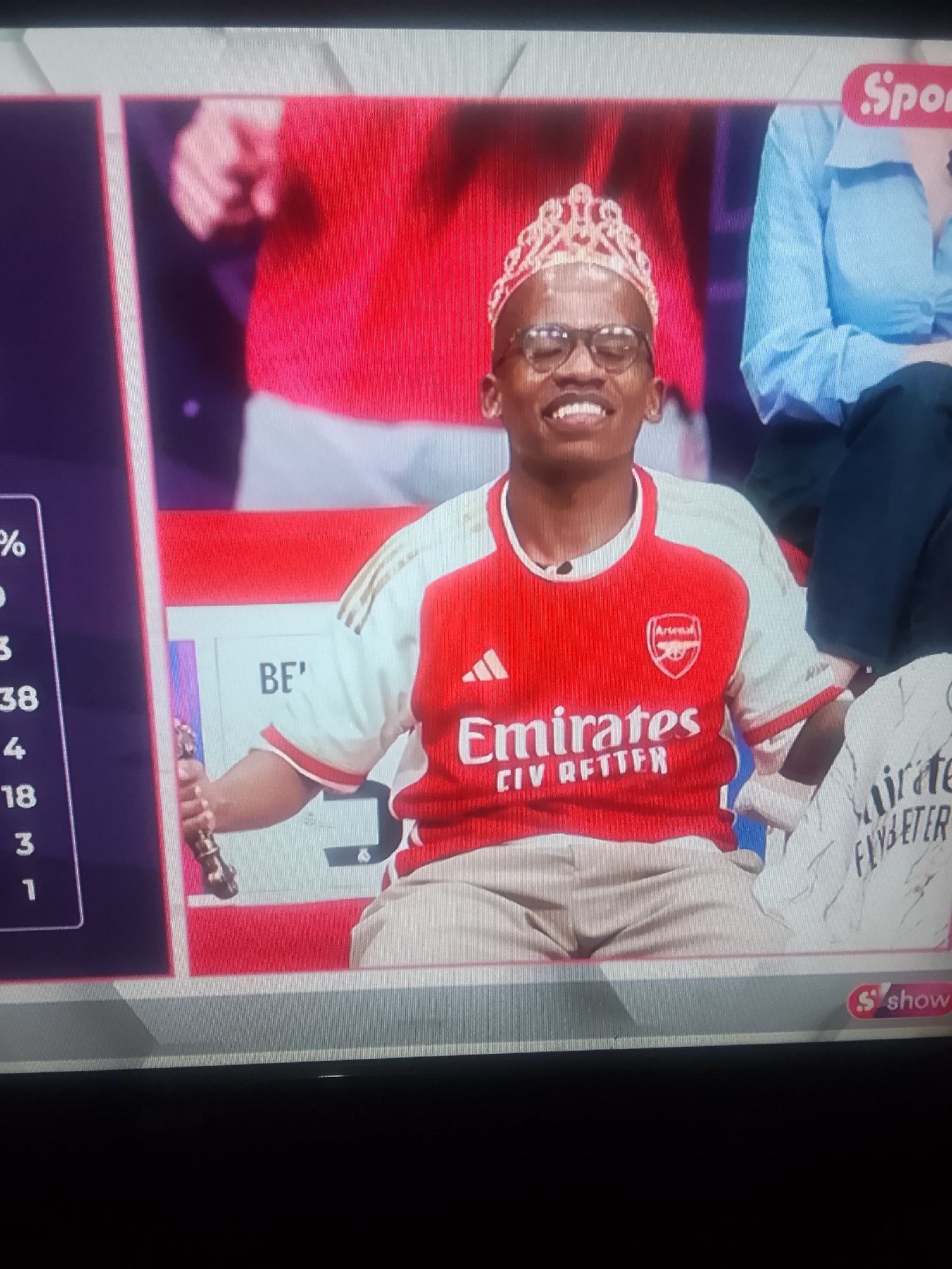 The image shows a man in an Arsenal jersey and a tiara, sitting in a studio setting. He is wearing glasses and smiling. To his left is a screen displaying percentages and the text "BE'". The Arsenal logo is visible on his jersey, along with the sponsor "Emirates FLY BETTER." To his right is a person wearing a blue shirt. The "Sport" logo and a red "S show" are visible in the upper and lower right corners, respectively. The city of Unknown, Unknown is not depicted in the image.