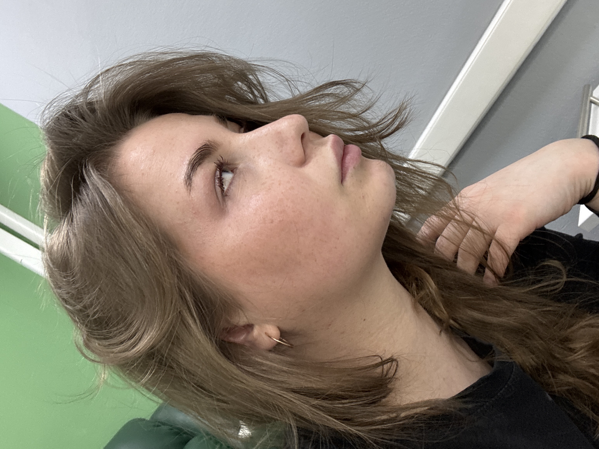 A low-angle, close-up shot of a woman with light brown hair, light skin with freckles, and a single gold hoop earring in her left ear. She is wearing a black shirt. Her head is tilted upwards and to the left, and her eyes are looking up. Her right hand is raised near her neck. The background shows sections of pale green and light grey walls, plus a white horizontal trim. It is not possible to ascertain the location as Chaikovsky, Russia from these visual cues.