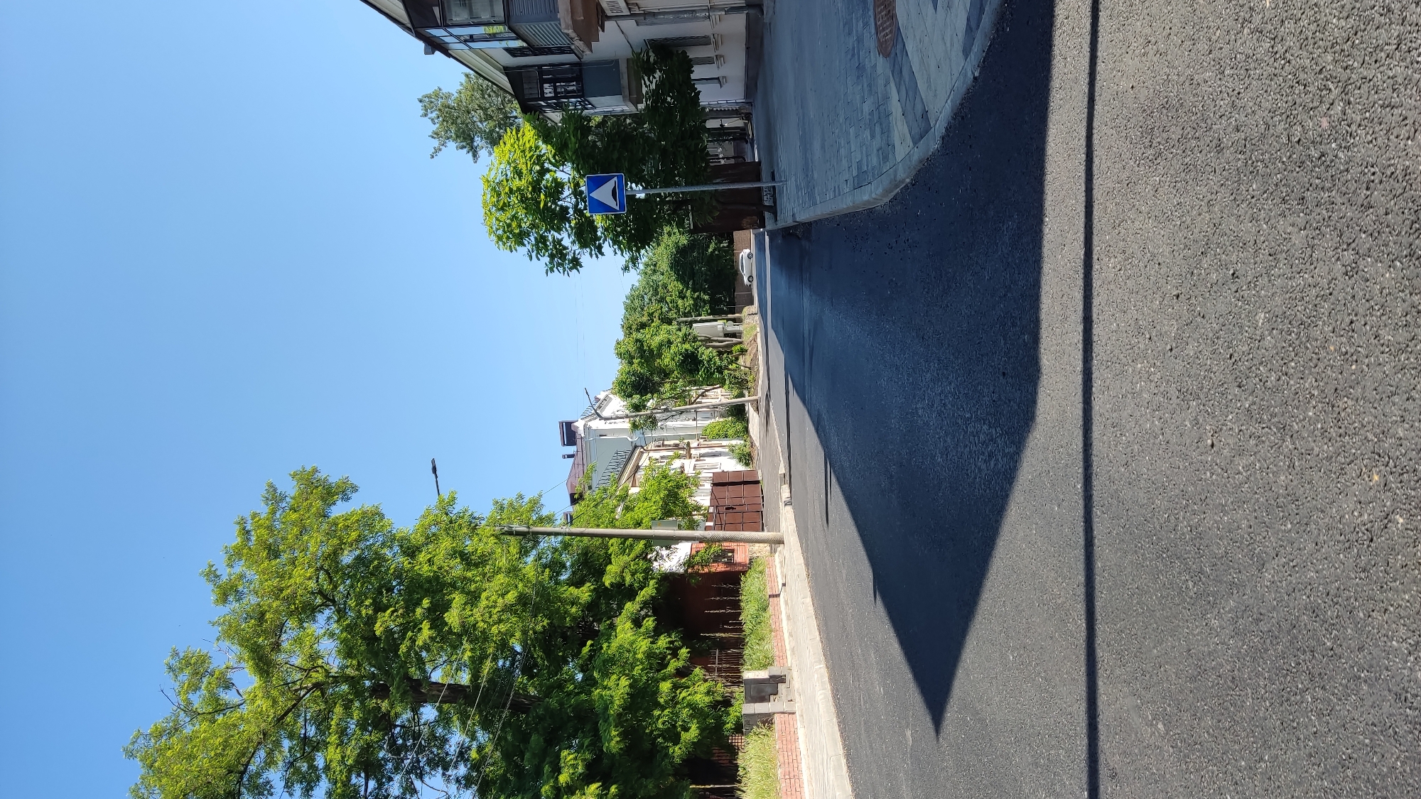 This is a street scene captured on a clear, sunny day in the city of Krasnodar, Russia. The image depicts a paved road, likely recently resurfaced with dark asphalt, stretching straight into the distance. A long, dark, geometric shadow extends across the foreground and midground of the road from the left.
On the left side of the street, a narrow concrete sidewalk runs alongside a low brick wall and a fence, behind which lush green trees and shrubs are visible. Further back, a white multi-story building with a brown roof is partially visible through the foliage, and a brown metal gate is situated at the entrance to a property. A utility pole stands prominently on this side, with a streetlight attached.
On the right side, a wider sidewalk paved with light grey interlocking pavers runs alongside another white multi-story building featuring dark window frames and an enclosed balcony on its upper level. A sign with Cyrillic text, reading "БАНЯ" (Bathhouse), is affixed to this building. A blue square road sign with a white triangular symbol indicating a speed hump is mounted on a pole near the right sidewalk. Abundant green trees line this side of the street as well.
In the distance, a white car is faintly visible on the street, and the road appears to curve slightly to the left beyond the visible buildings. The sky is clear and bright blue. No people or animals are present in the scene, and no specific actions or interactions are depicted.