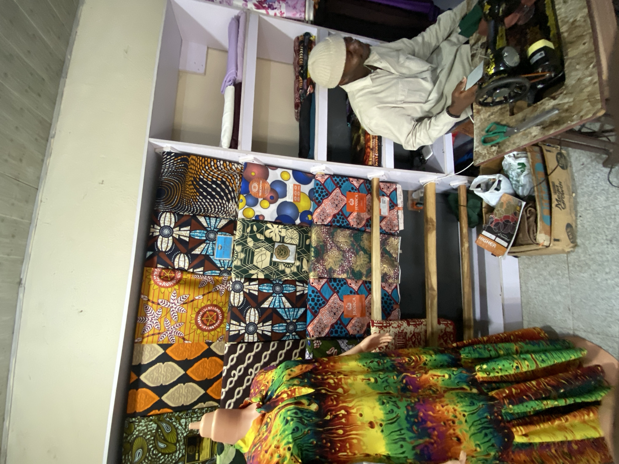 The image shows the interior of a fabric store in Bukuru, Nigeria. A man wearing a cream-colored traditional outfit and a white cap is sitting at a sewing machine, looking at a smartphone. A shelf is filled with brightly colored African wax print fabrics folded into rectangular shapes. A mannequin is partially visible at the bottom of the shelf, draped with more fabric. Under the sewing machine table, there's a cardboard box and plastic bags. The store appears to be compact and well-stocked with a variety of textiles.