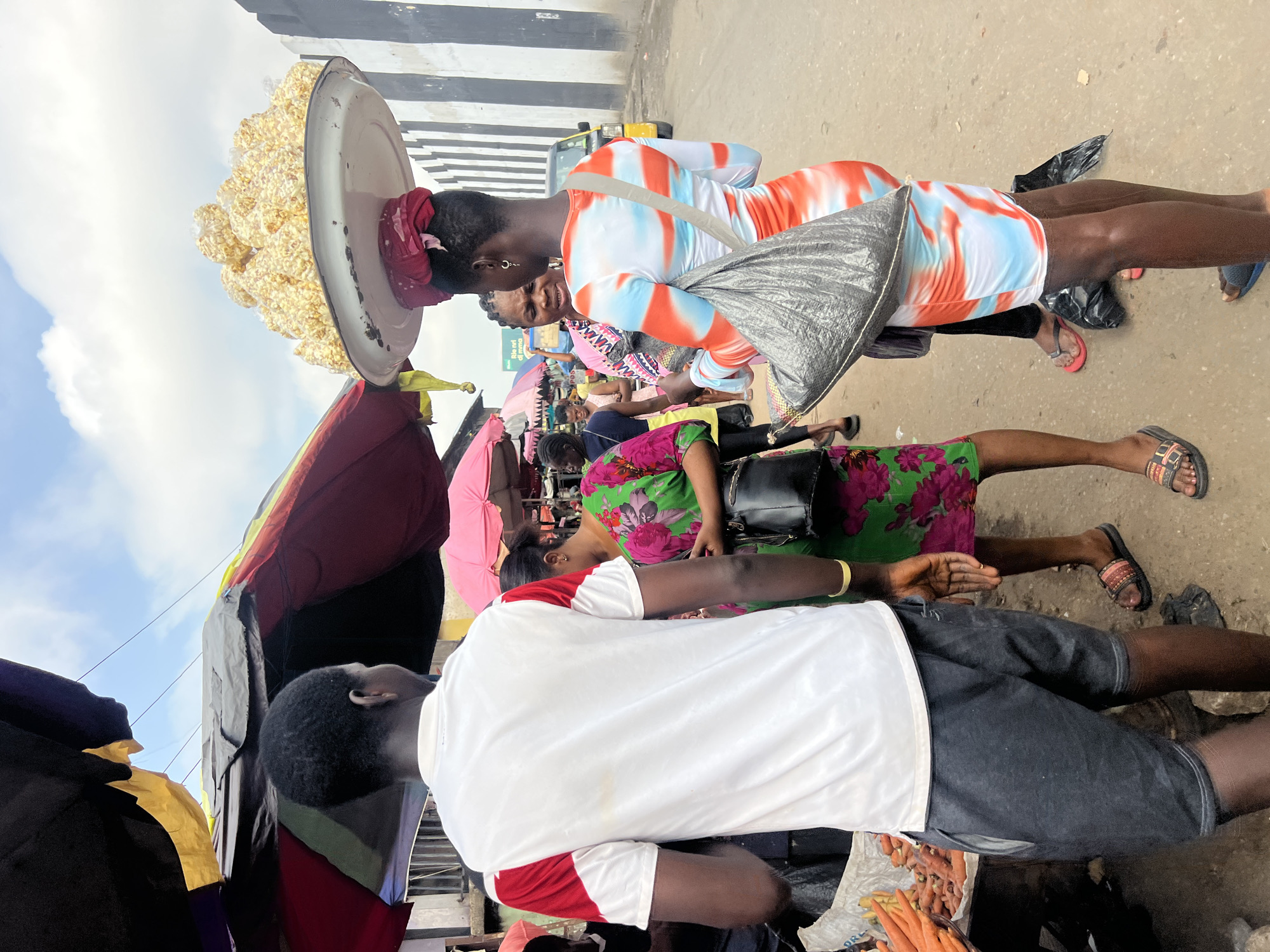 An outdoor market scene in Nkpor, Nigeria, features several people interacting. In the foreground, a man wearing a white t-shirt with red sleeves and dark shorts stands with his back to the camera, facing a stall piled with carrots. To his right, a woman in a green and pink floral dress holds a black handbag. Further right, another woman stands, wearing a tie-dye dress in orange, white, and blue. She has a large silver bag slung over her shoulder and a tray balanced on her head, filled with bags of popcorn. Behind her, other individuals and market stalls are visible, along with a yellow vehicle and a black and white striped wall. The sky above is partly cloudy.