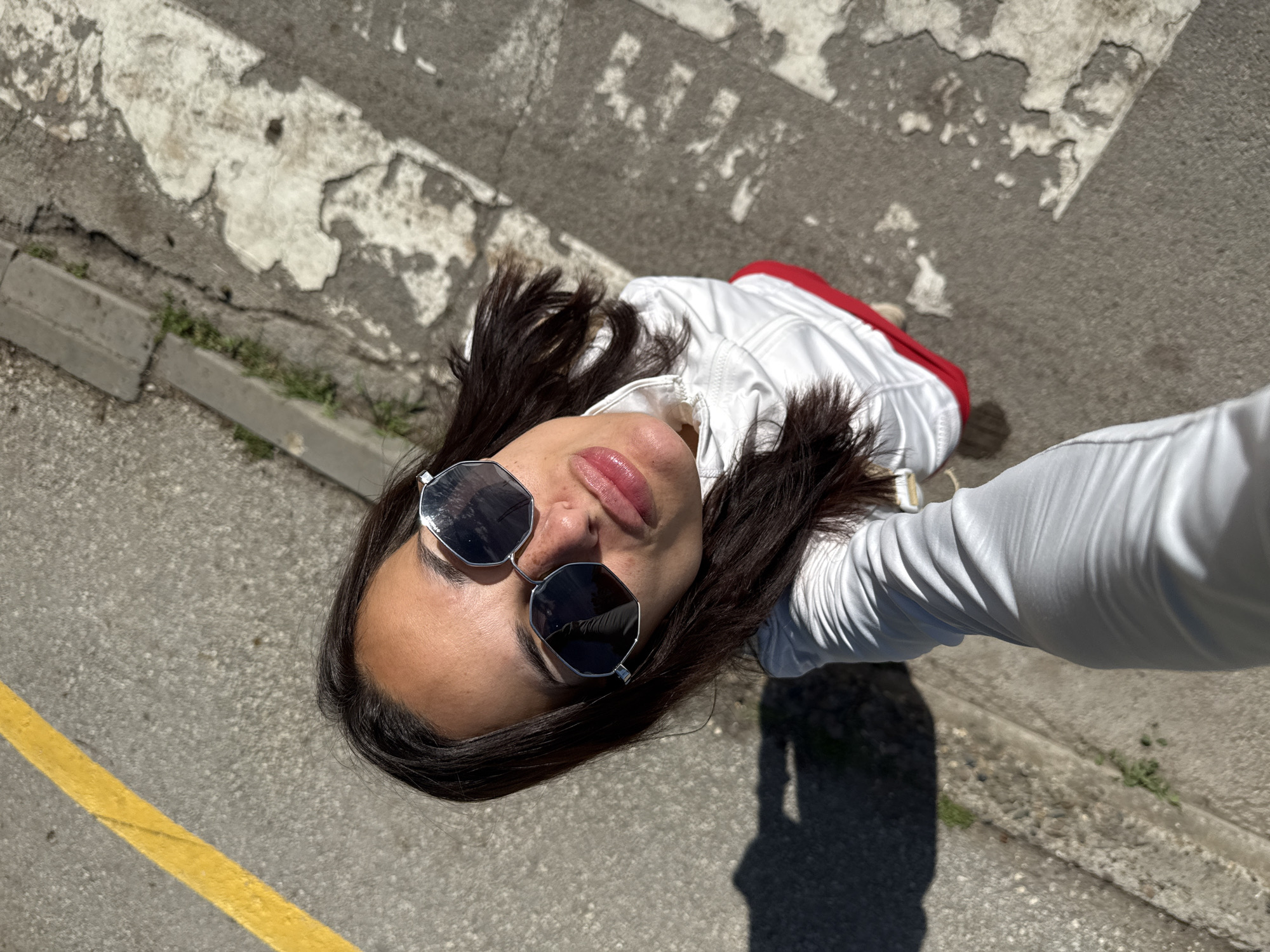 A woman is taking a selfie outdoors, possibly in Pleven, Bulgaria. She is wearing sunglasses, a white jacket, and red shorts. The background appears to be a street with a yellow line and a crosswalk. The lighting suggests it is daytime and sunny. The camera angle is looking down from above.