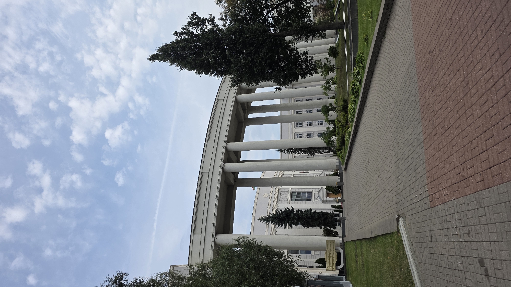 The image captures the National Academy of Sciences of Belarus in Minsk. A grand facade of the building is visible, characterized by a series of tall, white, fluted columns forming a covered walkway. The building itself is light-colored with numerous windows and appears to be several stories high. In front of the building, a paved walkway made of reddish-brown and grey bricks leads towards it. To the left and right of the walkway are patches of green grass, with mature trees, including tall evergreens and deciduous trees, dotting the landscape. A signpost with text in Cyrillic and English is partially visible to the left of the entrance. The sky above is light blue with scattered white clouds. The scene is static, with no people or animals present.
