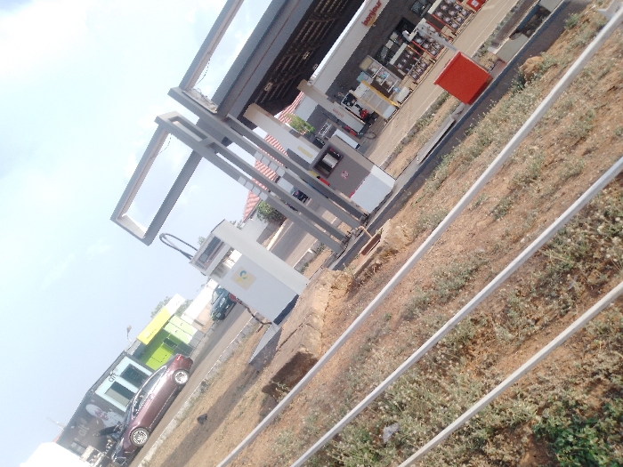 The image presents a ground-level view of a gas station in Sabon Tasha, Nigeria. Three parallel light-colored metal bars partially obscure the unpaved, reddish-brown foreground, which is sparsely covered with green vegetation. The gas station features a prominent dark-grey canopy supported by steel frames, extending over at least two white fuel dispensers. One dispenser displays a yellow and green circular logo. A dark-colored sedan is parked to the left of the pump area, and a dark-green vehicle is partially visible behind one of the pumps. Directly behind the pumps, a convenience store building is visible with "bonjour" signage and merchandise displayed through its windows. A red rectangular bin is situated near the store. Further in the background, a row of additional commercial buildings with various colored facades (yellow, green) can be discerned. The upper left portion of the scene shows a clear, bright blue sky. No individuals are clearly identifiable or performing specific actions within the frame.