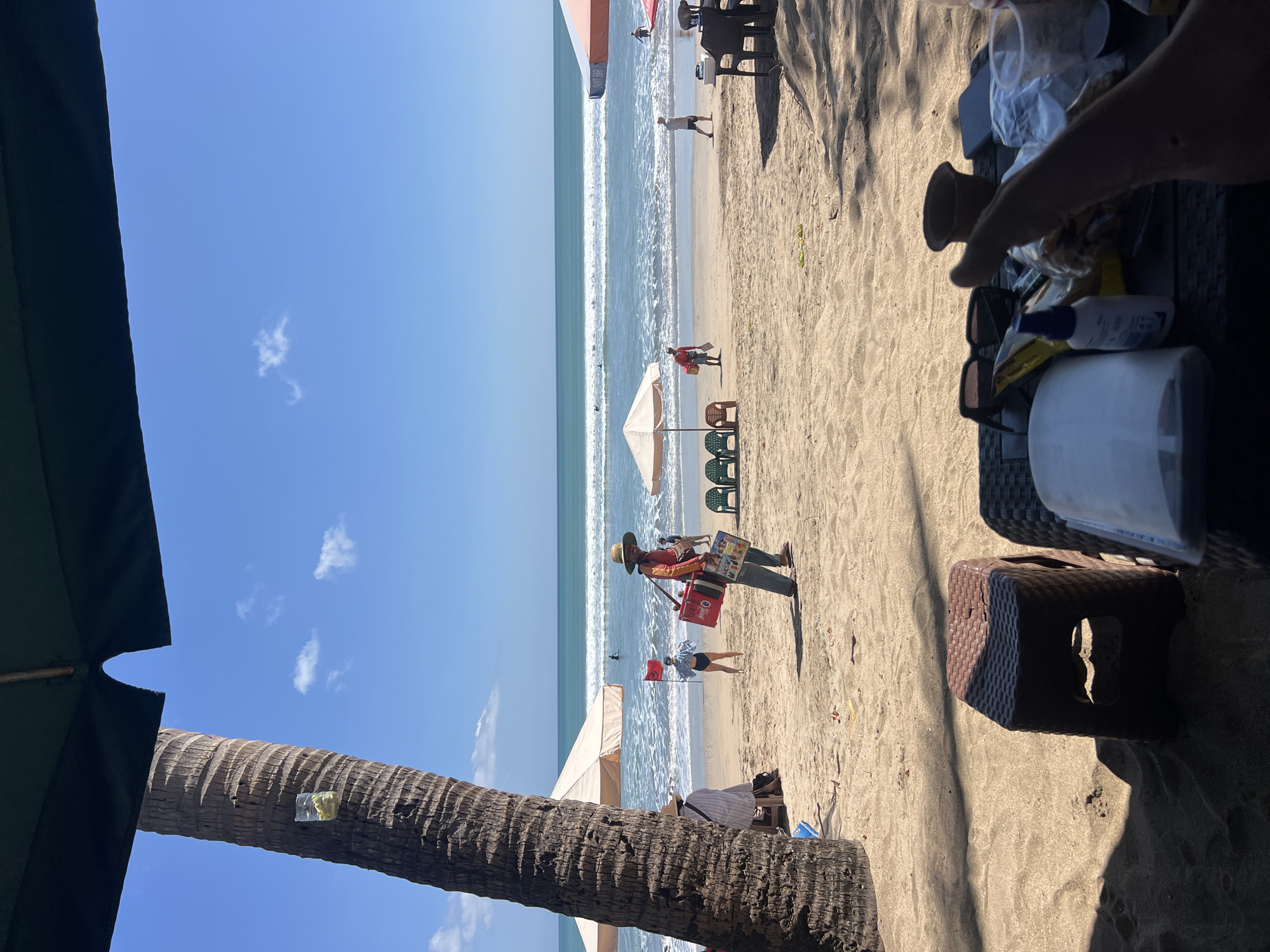 This is a high-angle, full shot of a beach scene in Kuta, Indonesia, on a sunny day. A palm tree dominates the left foreground, its trunk textured and scarred, with a clear plastic cup containing a yellow substance attached to it. Underneath a dark green umbrella, a table is set with a variety of items, including sunglasses, a small spray bottle, a plastic cup, a phone, and a brown pot. In the middle ground, a vendor wearing a hat and a red and yellow shirt walks across the sand carrying two coolers, one red and one white, on his shoulders. He is holding a red flag. Other beachgoers are scattered across the sand, some sitting under umbrellas and others near the water. In the distance, the turquoise ocean stretches to the horizon, with gentle waves breaking on the shore and a few surfers visible in the water. The sky is a clear blue with a few wispy white clouds. The sand is light brown and appears to be fine-grained.