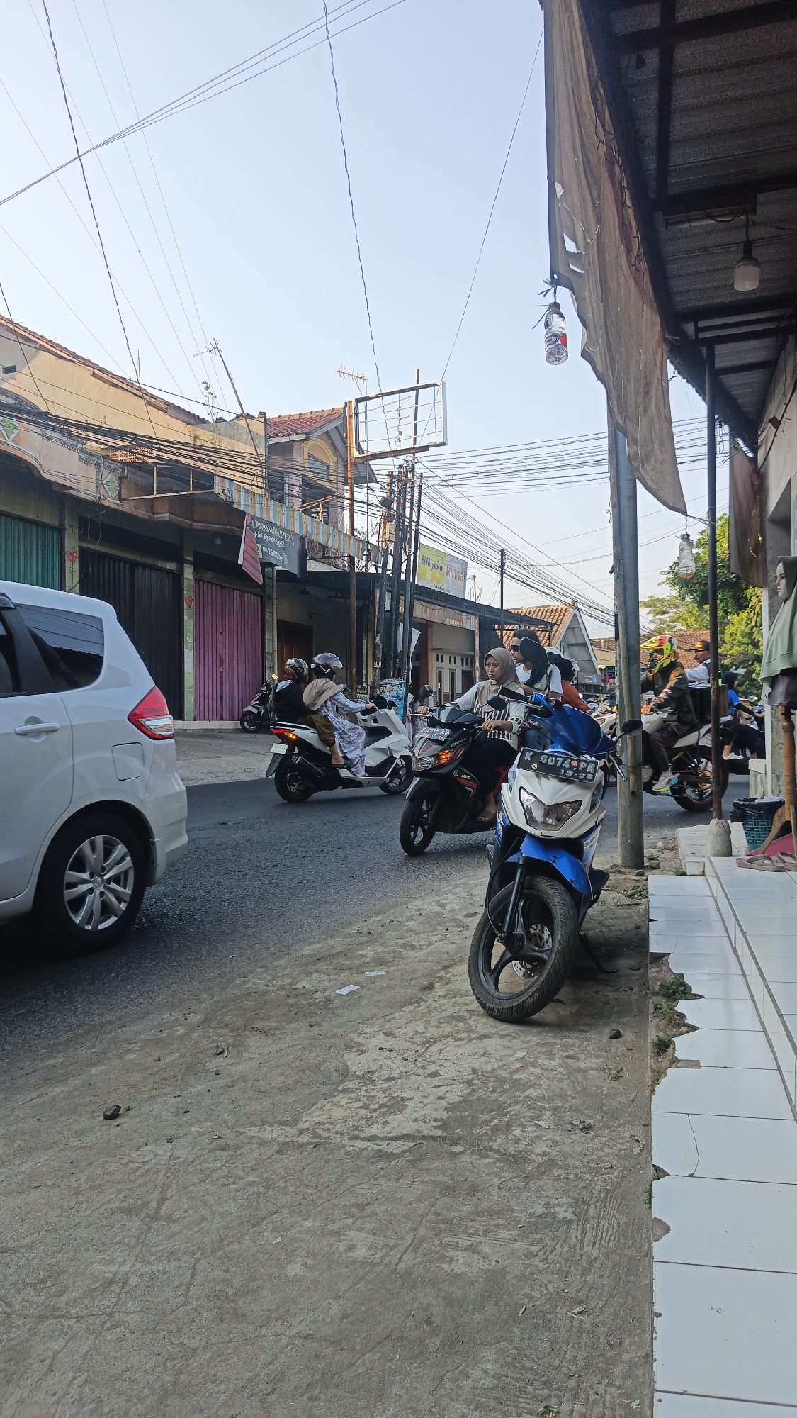 The image depicts a street scene in Dawe, Indonesia, likely during daylight hours given the clear sky and sunlight. The perspective is from a low angle, looking down a street lined with buildings on both sides.
On the left side of the street, a white car is partially visible in the foreground. Behind it, several buildings with varying architectural styles and colors are present. Some have closed storefronts with pink and green doors, while others appear to be residential. A complex network of electrical wires crisscrosses above the street and buildings.
The right side of the street features a sidewalk with white tiled paving, some of which is damaged. A covered awning extends from the buildings, with a tattered tarpaulin hanging from it. A light fixture is suspended from the awning. In front of the buildings, a row of parked motorcycles is visible.
Several people are present, primarily riding motorcycles. In the middle ground, two women wearing hijabs are riding on a white scooter, with one driving and the other as a passenger. Further down the street, more individuals are seen on motorcycles, some wearing helmets. In the foreground on the right, a blue and white motorcycle is parked. A mannequin dressed in green is visible inside a shop on the far right. The road surface appears to be asphalt with some patches and debris. The overall impression is of a busy, everyday street in a populated urban area.