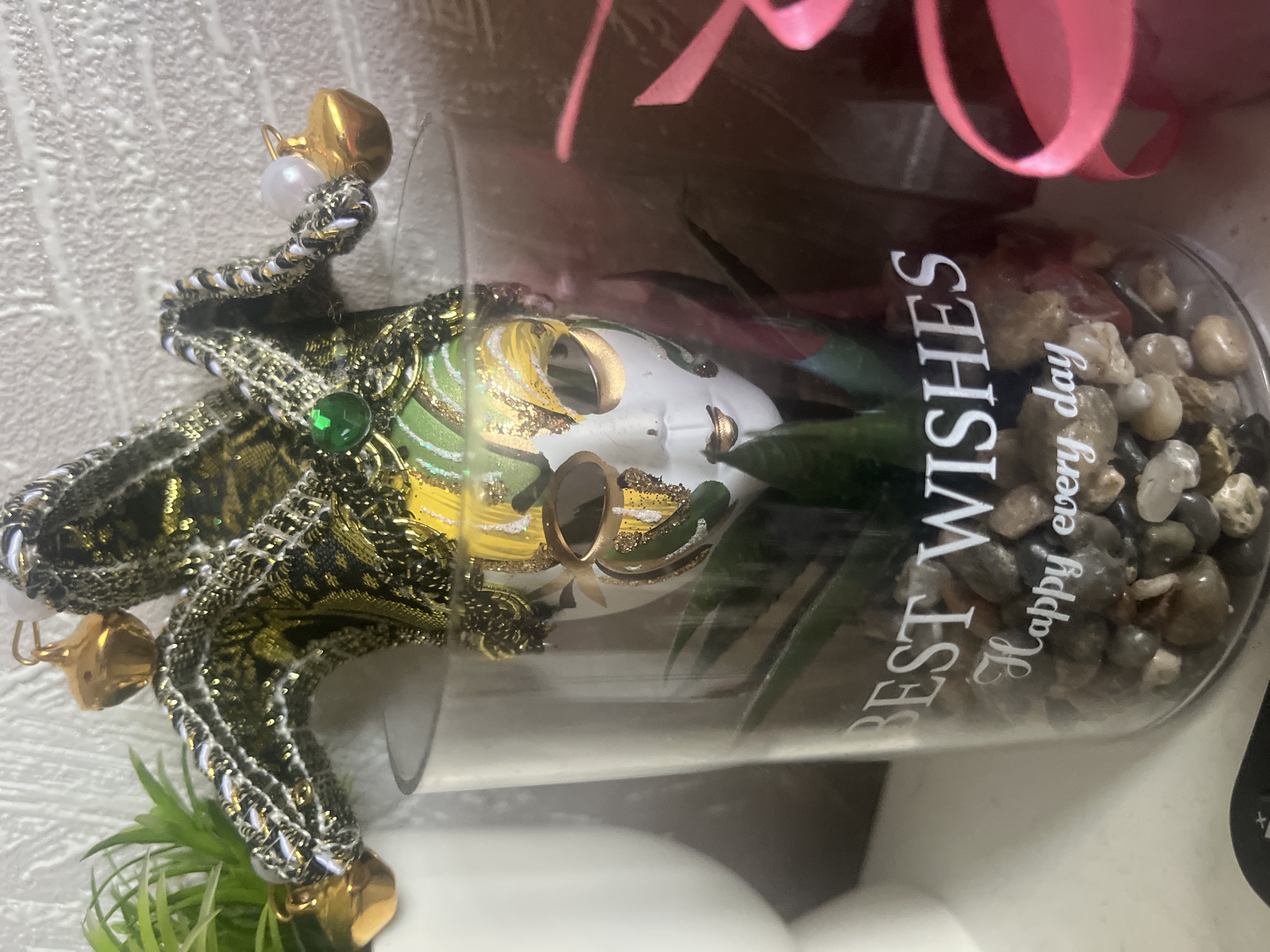 A close-up shot presents a decorative item consisting of a jester mask atop a glass container. The mask features a black and gold patterned fabric, adorned with small gold bells, a green gem, and faux pearls. Inside the glass container are small rocks and green plants, with the words "BEST WISHES Happy every day" printed on its surface. A pink ribbon is visible in the background. The location is not discernible from the image.
