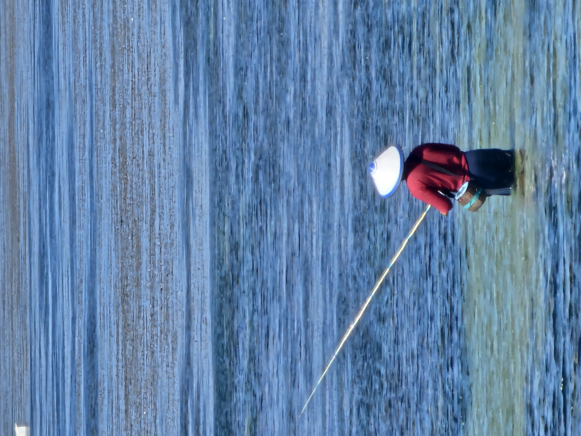 A person wearing a conical hat, red shirt, and dark pants stands in shallow, clear blue water, holding a fishing rod. A woven basket is slung over their shoulder. The water exhibits subtle ripples and variations in color, suggesting a shallow seabed beneath. The person appears to be fishing, with the rod extended outwards. The background consists of more blue water, with horizontal lines indicating movement or texture on the surface. This scene is likely located in a coastal area of Denpasar, Indonesia, perhaps near Sanur or Kuta beach, where shallow waters are common for recreational fishing.
