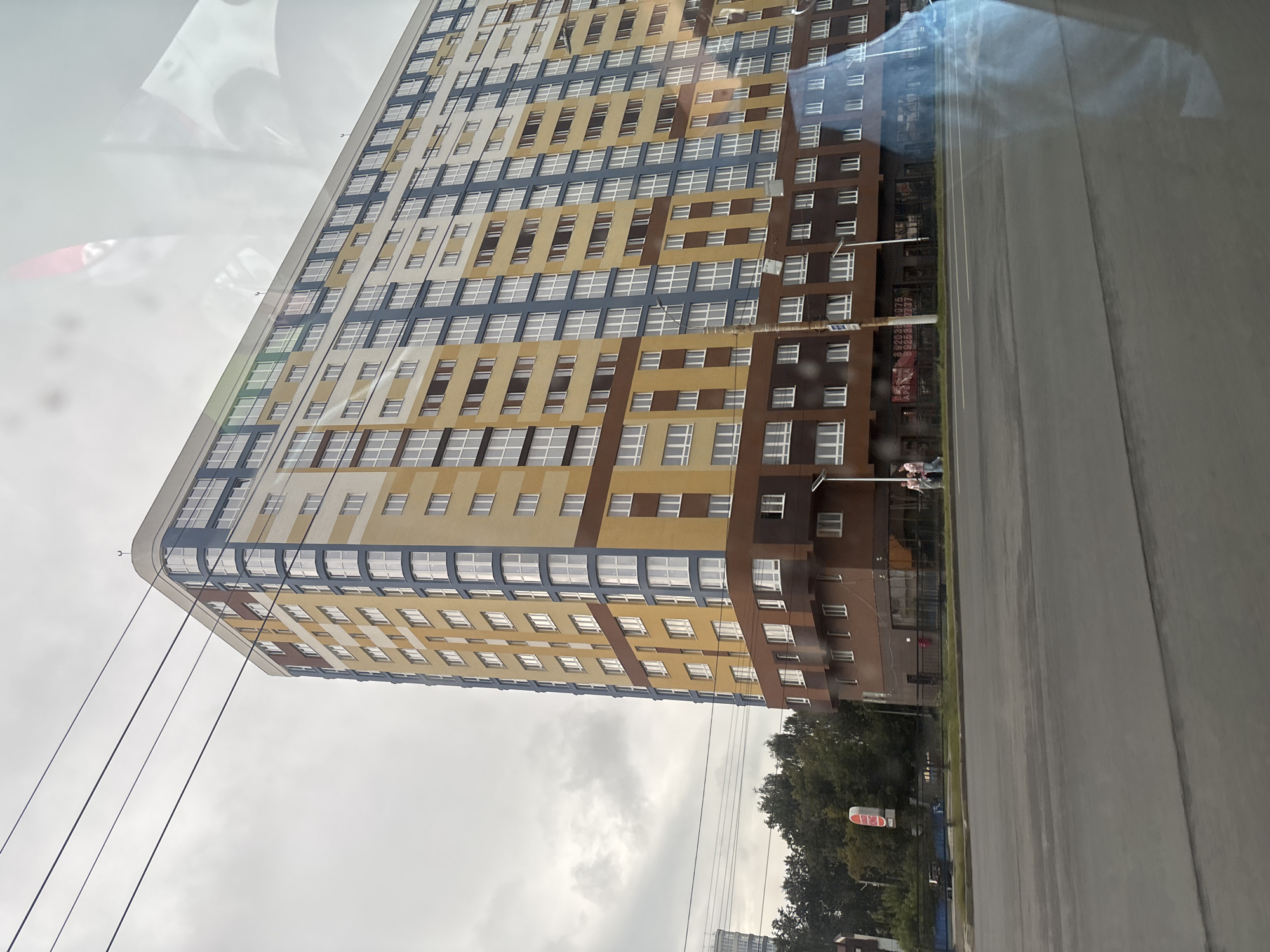The image depicts a modern, multi-story residential building in Ivanovo, Russia. The building is characterized by its colorful facade, featuring sections of yellow, blue, and brown. Numerous windows are visible across the building's exterior. In the foreground, a paved road runs horizontally across the image, with white lane markings. Several utility poles and wires are present, with the wires stretching diagonally across the sky. Trees and other smaller buildings can be seen in the background. The sky is overcast with clouds. Reflections are visible on the surface of what appears to be a window, obscuring parts of the scene. Two individuals are standing on the sidewalk in front of the building.