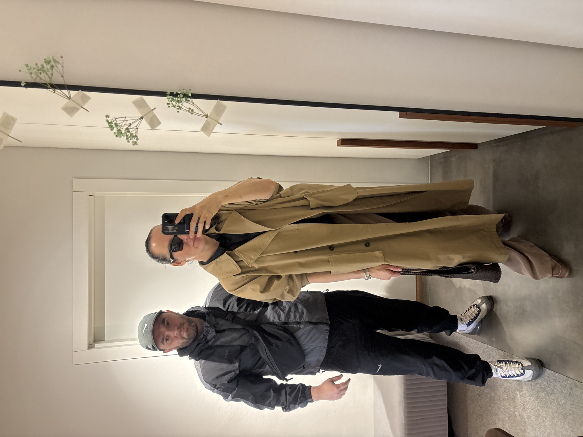 A man and a woman are taking a selfie in a mirror in what appears to be a store or showroom. The woman, at the top of the frame, is holding a smartphone and wearing sunglasses and a tan trench coat. The man, below her, is wearing a gray and black jacket with a black bag, black pants with a white Nike logo, and gray and white sneakers. He also wears a gray baseball cap.
The background includes a white wall with small floral decorations attached with tape, a brown wooden frame, and a gray concrete floor. A white door is visible to the left of the man. There is no definitive evidence to suggest this image was taken in Chaikovsky, Russia.