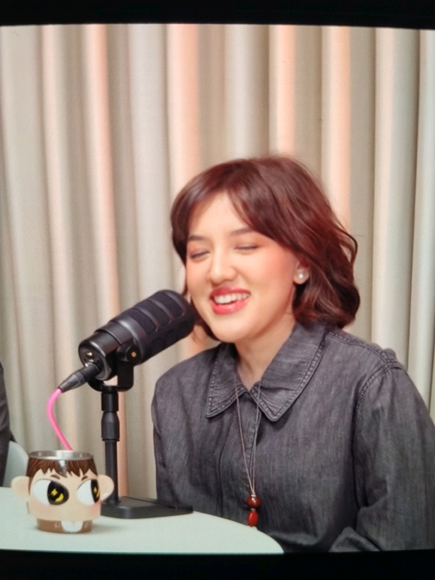 A woman with short, dark brown hair and fair skin is sitting at a table, smiling with her eyes closed. She is wearing a dark gray denim shirt and a pearl earring. A black microphone is positioned in front of her. To her left, on the table, is a novelty mug shaped like a cartoon character's head, with a pink straw extending from it towards the microphone. The background consists of pale, vertical curtains. The image appears to be a screenshot from a video, possibly a podcast recording, and the setting is likely a studio. The provided location of "Korleko Selatan, Indonesia" cannot be verified from the visual information in the image.