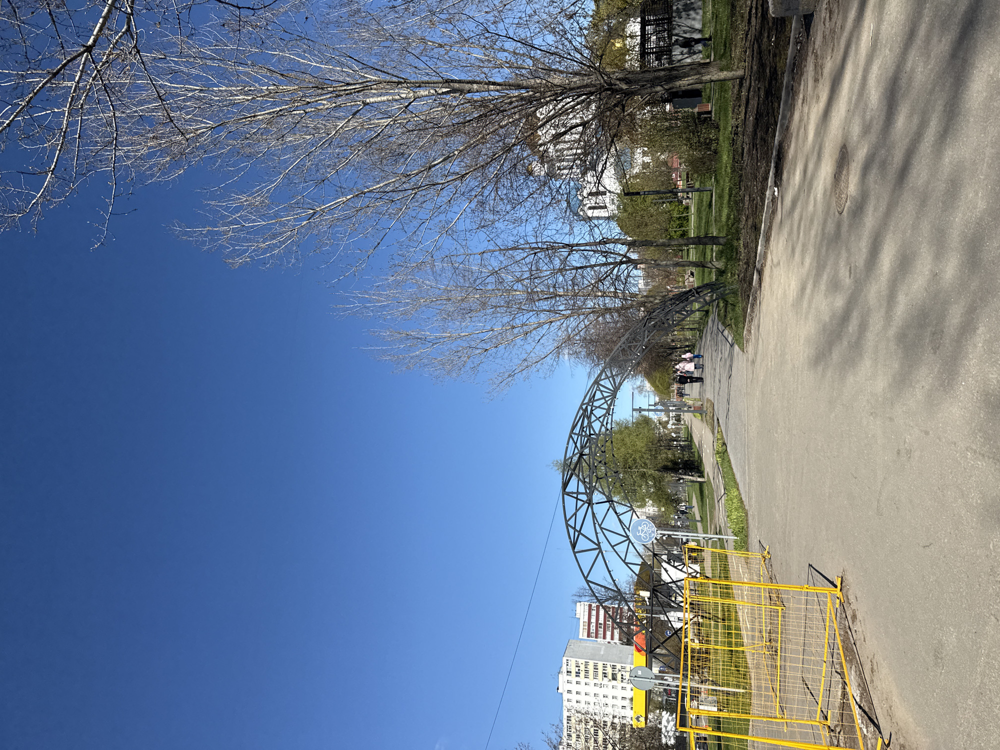 The image depicts a street scene in Moscow, Russia, characterized by a wide, concrete sidewalk. Adjacent to the sidewalk is a green strip with trees, some still bare, suggesting early spring. A notable architectural structure, possibly a decorative arch or support, is present. Yellow metal barriers are situated along the sidewalk. In the background, multi-story residential buildings are visible under a clear blue sky. The photograph is taken at a low angle. There are people walking in the distance on the sidewalk.