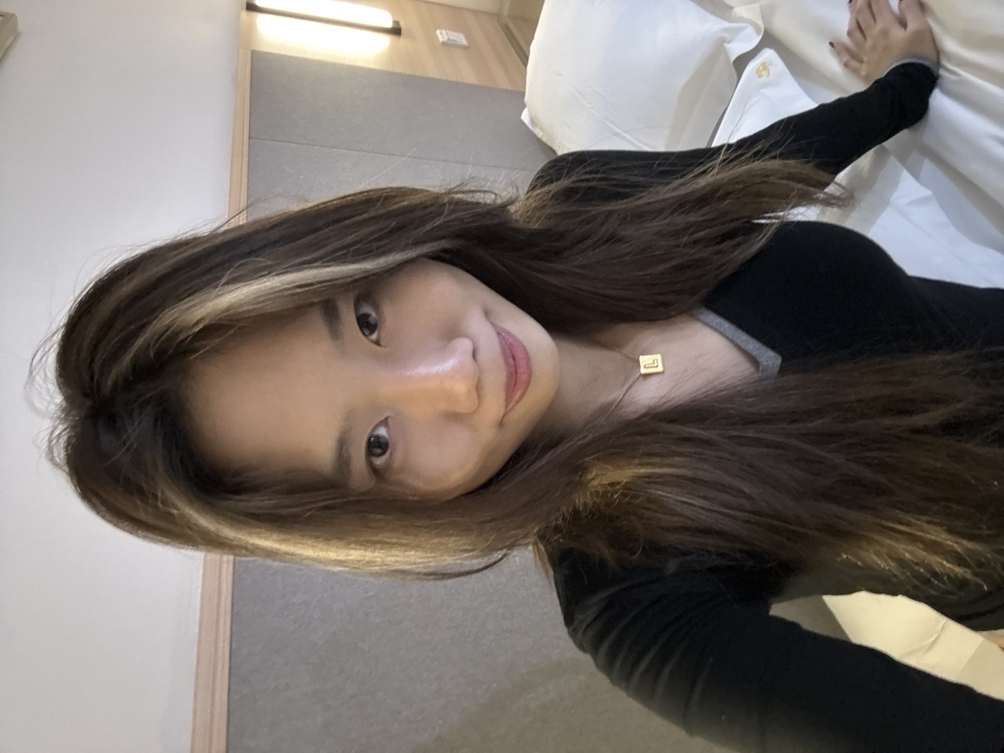 A woman with long, brown hair is lying on a white bed in a hotel room. She is wearing a black, long-sleeved shirt and a gold necklace. The room has grey walls and a light fixture is visible near the top left corner. The location appears to be a hotel, but there is no direct indication of its location in the Yuzhong District of China within the image.