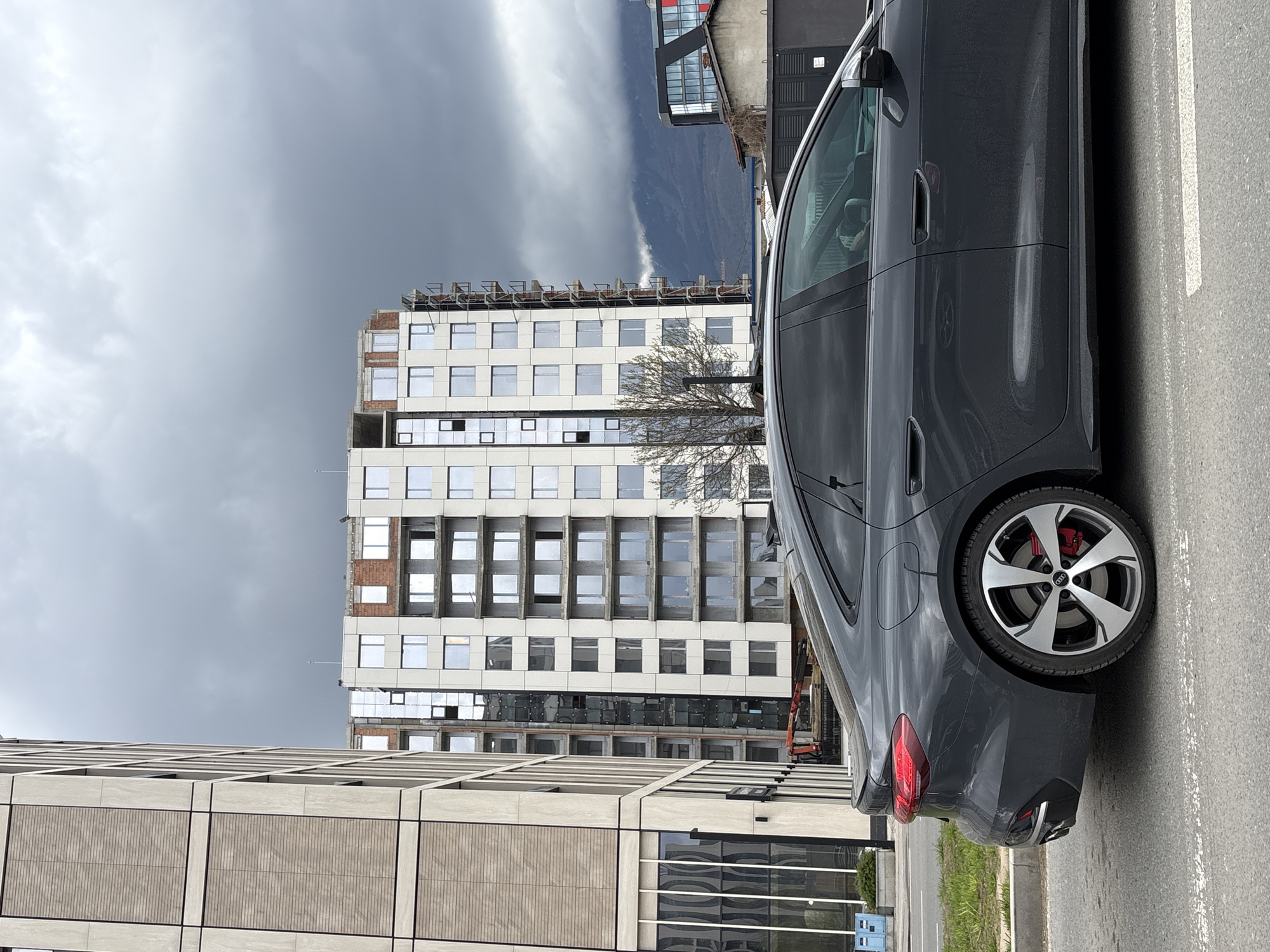 A medium, eye-level shot of a grey car parked in a parking spot along a street in Sofia, Bulgaria. The car's driver's side is in view, from the front door to the rear of the vehicle, angled slightly in relation to the viewer. The sky is grey with dark storm clouds. There are two large buildings in the background, both partially under construction. An orange construction crane is visible between the buildings. The building closer to the car is light tan with concrete-textured paneling. The taller building behind it is white with numerous square windows, some covered by reflective coverings. A bare tree stands between the buildings. Mountains are visible in the distance. A white parking line separates the car from the street. Green foliage lines the edge of the curb.