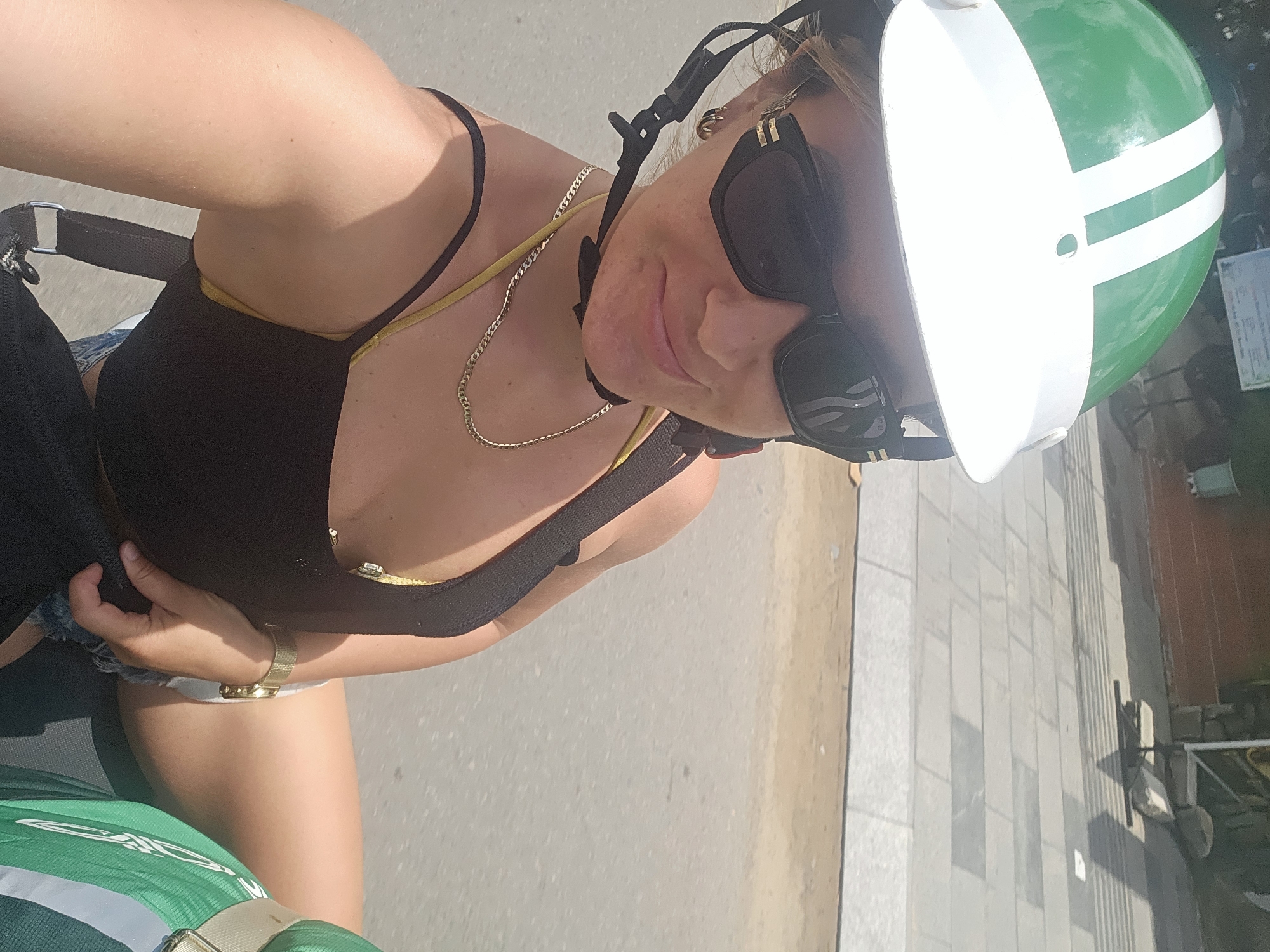A woman is riding a motorcycle in Phu Thuy Ward, Vietnam, and is taking a selfie. She is wearing a green and white helmet, sunglasses, a black tank top with yellow straps, and denim shorts. A gold chain necklace and a gold watch are visible. The background shows a paved sidewalk and road.