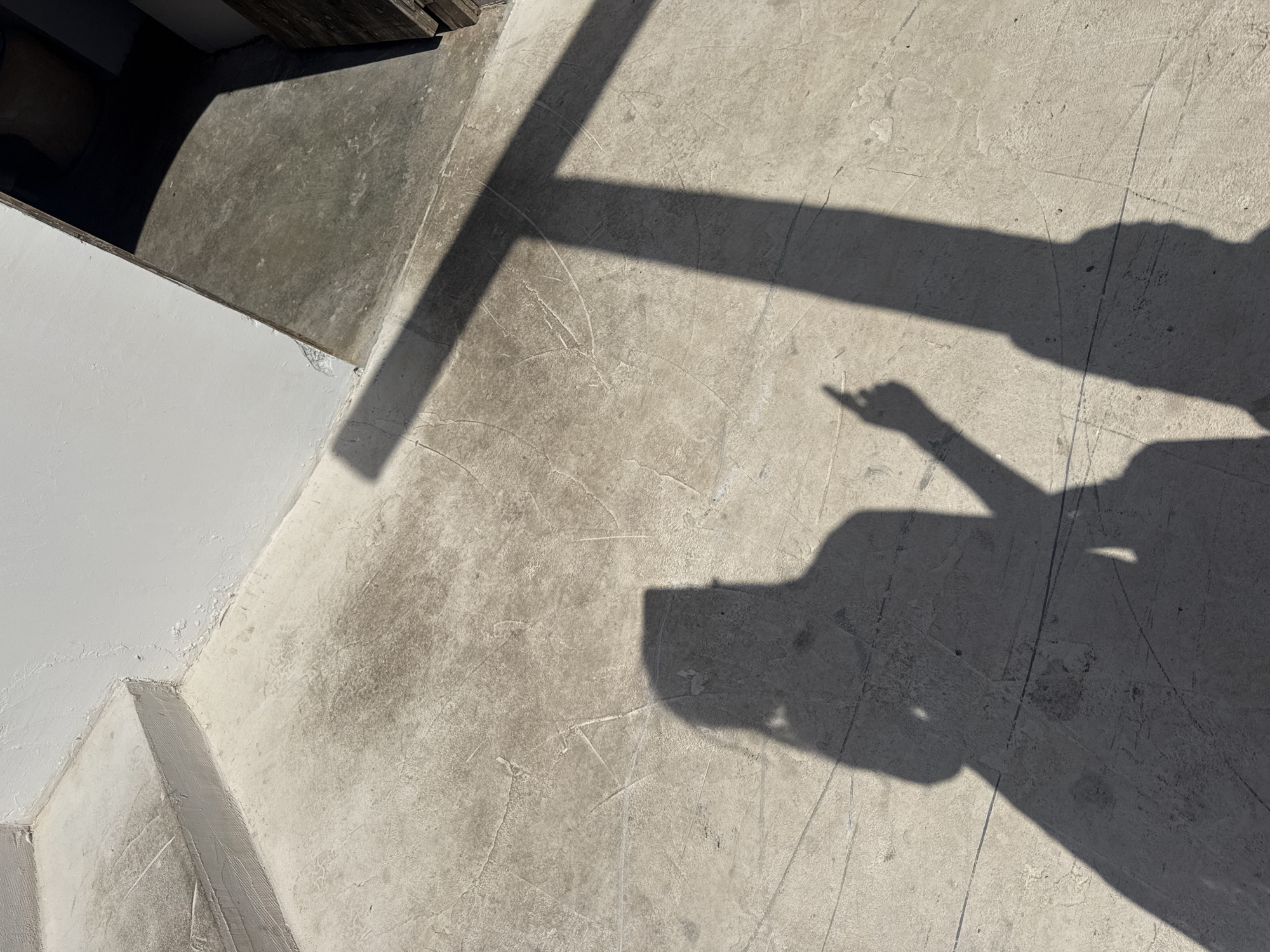 The image presents an outdoor scene dominated by concrete ground and prominent shadows. In the upper left corner, a white wall meets the concrete surface, adjacent to a dark, recessed area which could be a doorway or an interior space. The concrete ground is textured with numerous visible cracks and fine lines.
A large, dark, cross-shaped shadow, likely cast by a window frame, railing, or other architectural element, stretches across the upper and central portions of the ground. Overlapping this and to the right, are the distinct shadows of at least two individuals. One shadow shows a partial silhouette of a person's upper body, including the head and torso. Another, clearer shadow depicts an arm and a hand with an extended index finger, appearing to point towards something.
The sharp definition and darkness of the shadows indicate bright, direct sunlight, suggesting a clear and sunny day, most likely around midday or early afternoon when shadows are most pronounced. There are no discernible location cues beyond the concrete and white wall, and no visible text in the image.