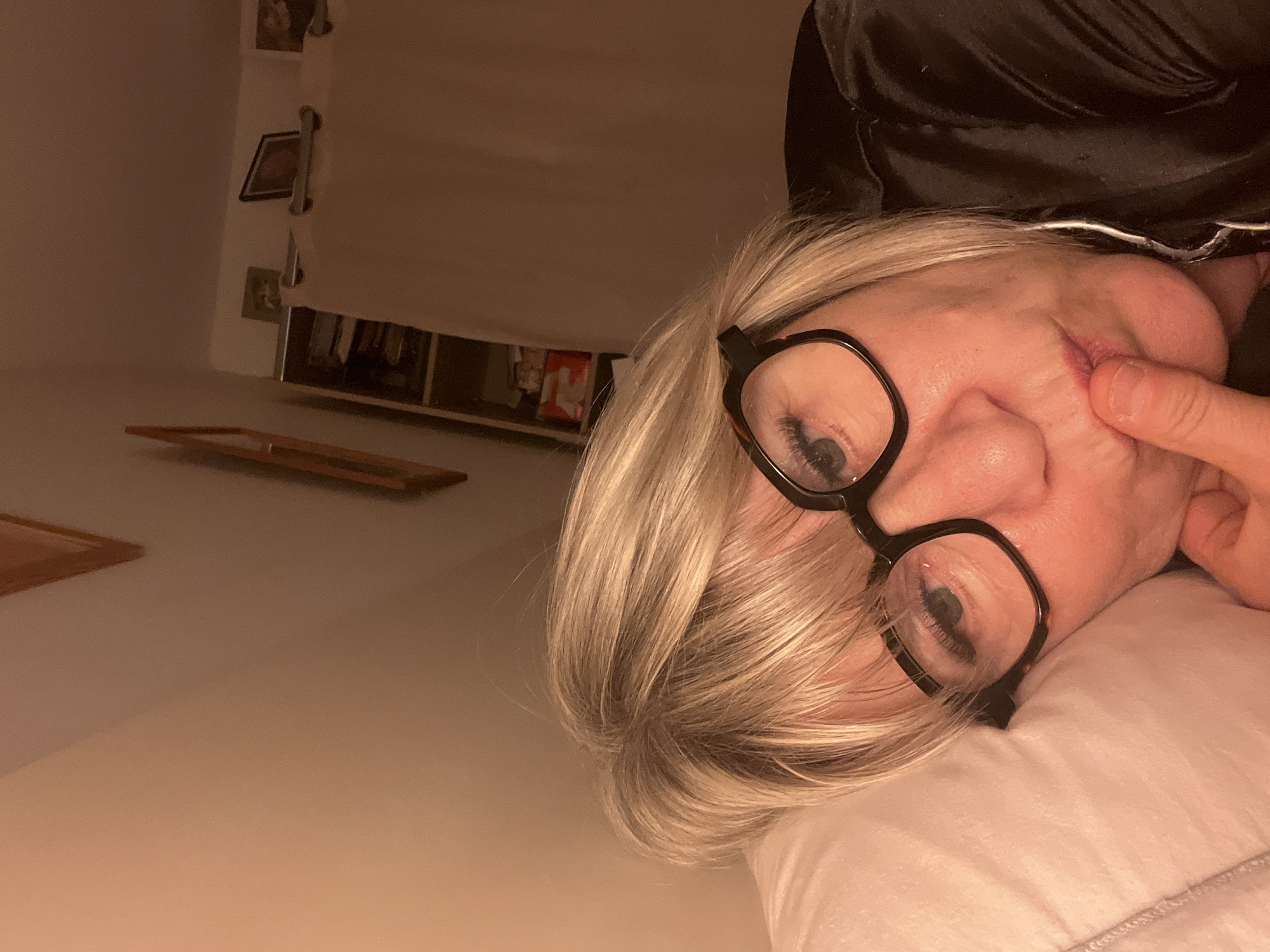 The image depicts a woman with blonde hair and black-rimmed glasses lying on a white pillow. She is wearing a black top, and her right index finger is touching her cheek. There appears to be a lesion on her face near her nostril. The background includes a wall, a window, and what appear to be picture frames. The location, Chambœuf, France, is based on additional context not present in the image itself and therefore could not be derived solely from its contents.