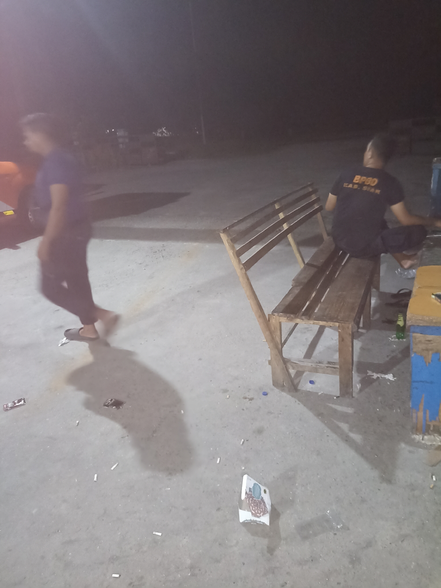 In Kampung Tengah, Indonesia, a man wearing a black t-shirt with "BP30 KAB. SIAK" printed on the back sits on a wooden bench. Next to the bench is a partially visible wooden table with blue paint on its side. A second person, blurred by motion and wearing a purple shirt and dark pants, walks by in the foreground. The ground is paved and littered with cigarette butts. The scene is dimly lit, suggesting it is nighttime.