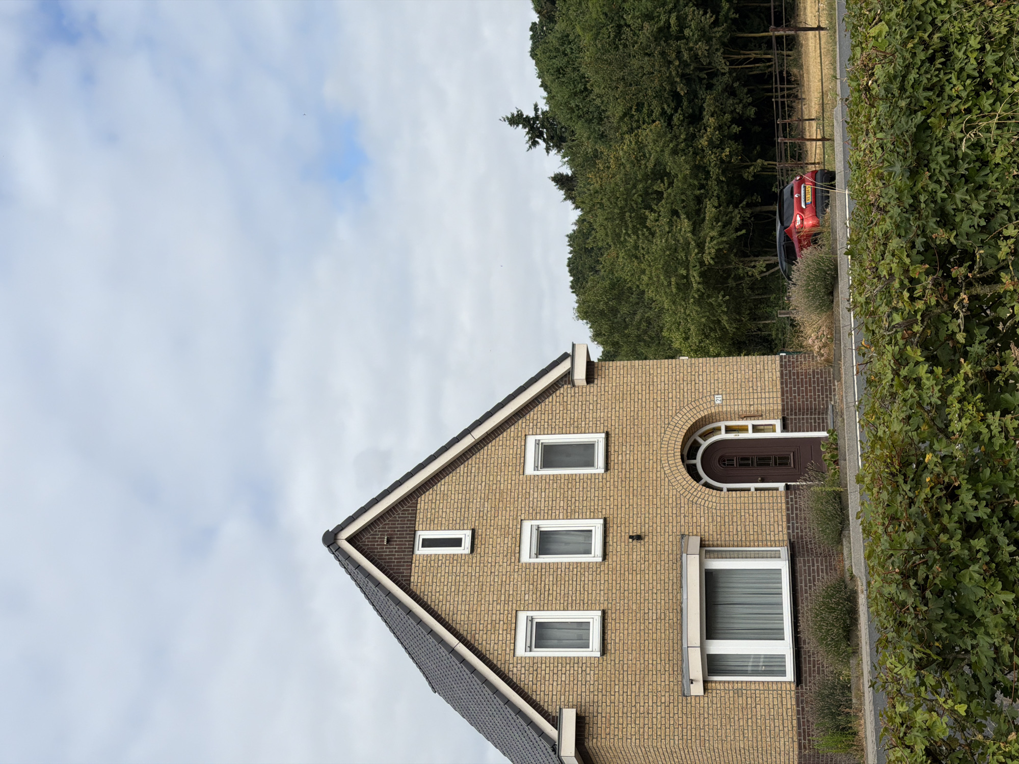The image shows a two-story brick house with a dark grey tiled roof. The house is situated in the city of Melick, Netherlands. The front of the house features several white-framed windows of varying sizes, with the largest window on the ground floor being a bay window. A dark brown arched door is visible on the ground floor, to the right of the bay window. The house is surrounded by greenery, with a dense hedge in the foreground and a treeline in the background. A red SUV with a Dutch license plate is parked on the right side of the image, beyond a fence. The sky is overcast with scattered clouds.