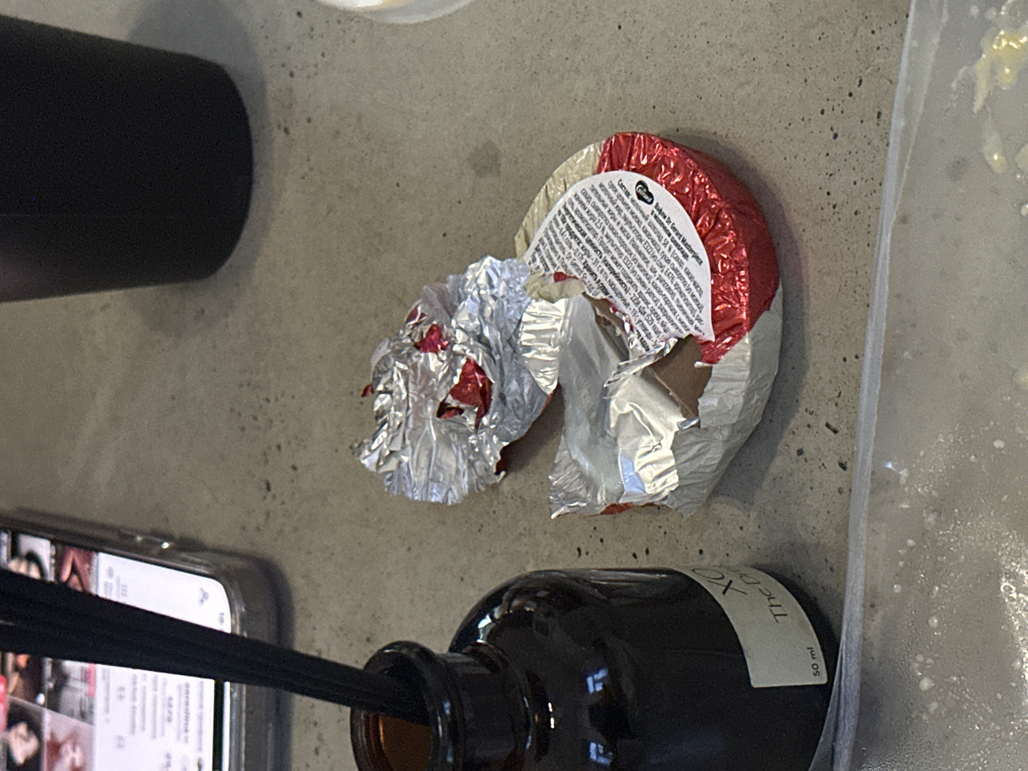 The image shows a still life of discarded items on a gray, possibly concrete, surface. A black cylindrical object is visible on the left. An opened, crumpled candy wrapper with red foil and a circular label with text in Russian is also visible. Below the wrapper is a brown glass bottle with a white label that reads "Theo" and "50 ml." The neck of the bottle contains black diffuser sticks. A smartphone is positioned near the bottle, displaying a screen with several images or articles. The edge of a clear plastic bag or container is visible on the right side of the image. The presence of Russian text suggests the location may be in Moscow, Russia.