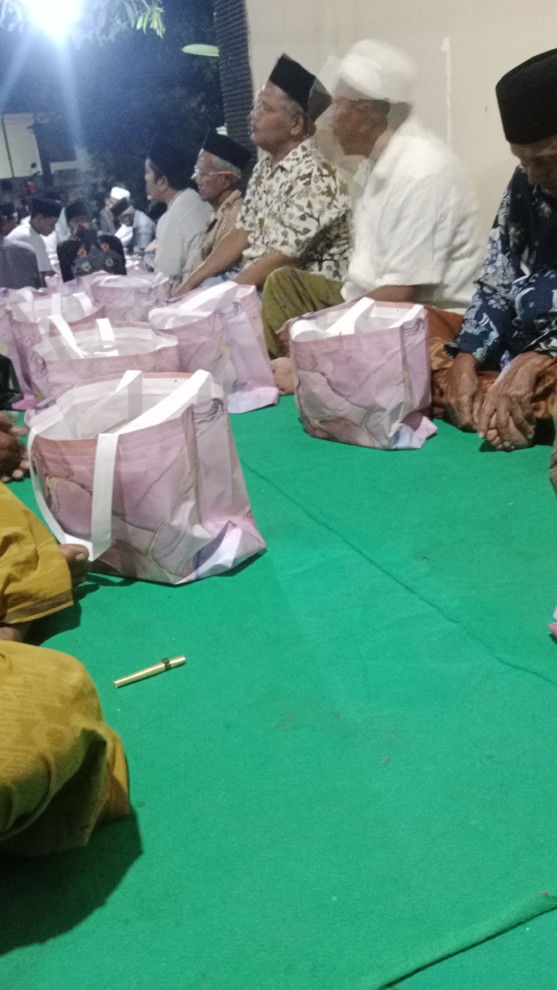 The image depicts an outdoor gathering in Pondok Kelor, Indonesia, likely a social event or religious ceremony given the attire of the participants. Several men, predominantly wearing traditional Islamic clothing such as peci (caps) and sarongs, are seated on a vibrant green carpeted surface. In the foreground, several patterned bags are arranged, possibly containing gifts or food. The lighting suggests it is evening, with artificial lights casting shadows. One person in the foreground is wearing a yellow garment. The background shows a building wall and trees, indicating a residential or community area.