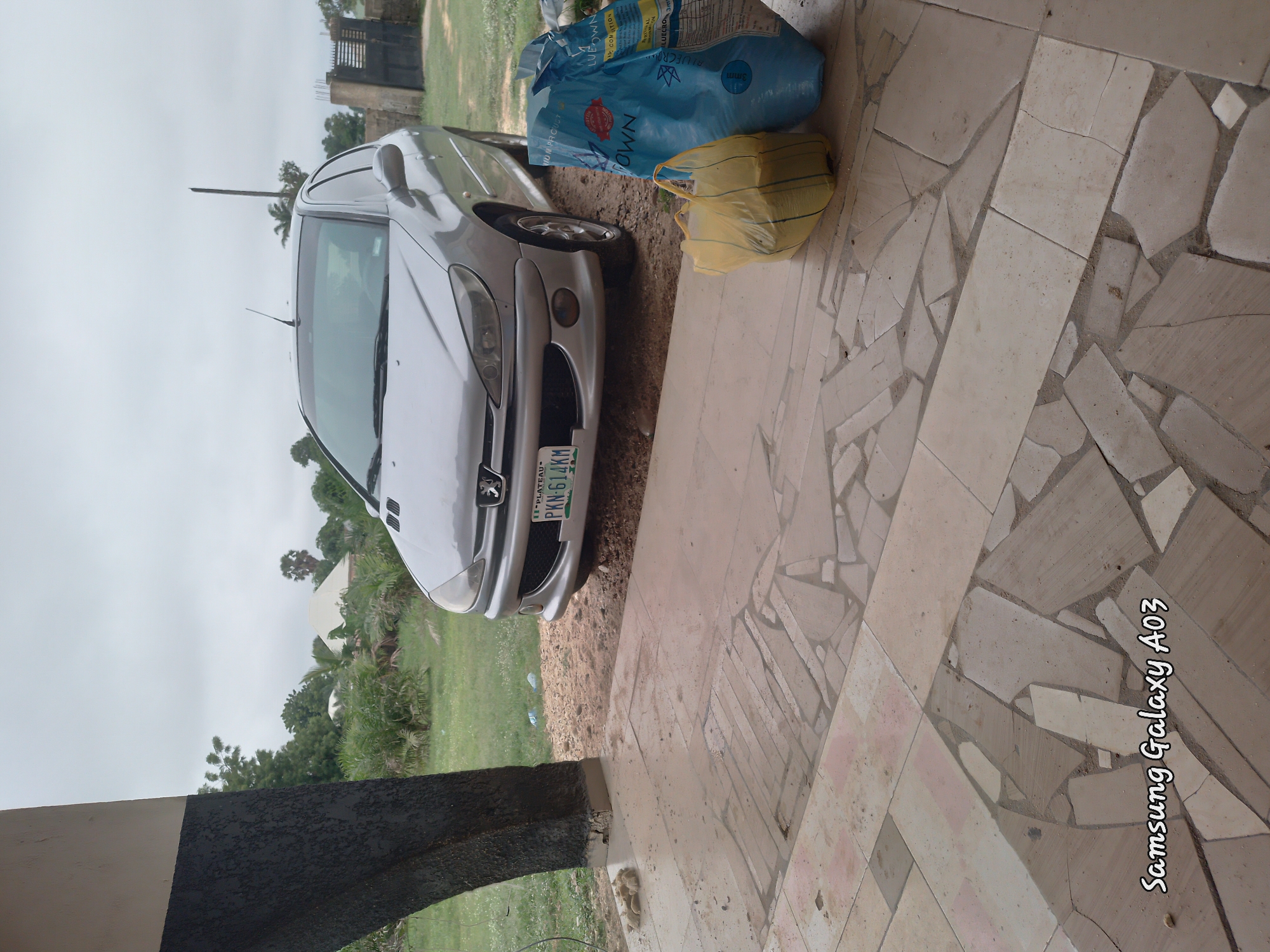 In Doka-Tofa, Nigeria, a silver Peugeot 206 sedan, license plate PKN-614KM, is parked on a dirt and gravel surface near a tiled patio. Two large bags, one blue and one yellow, are placed on the patio in front of the car. The scene is partially shaded by an overhead structure with a dark, textured pillar. In the background, lush green vegetation and trees surround some buildings, under an overcast sky. The tiled patio features a mosaic of irregularly shaped beige and brown tiles. The timestamp "Samsung Galaxy A03" is visible in the bottom left corner of the image.
