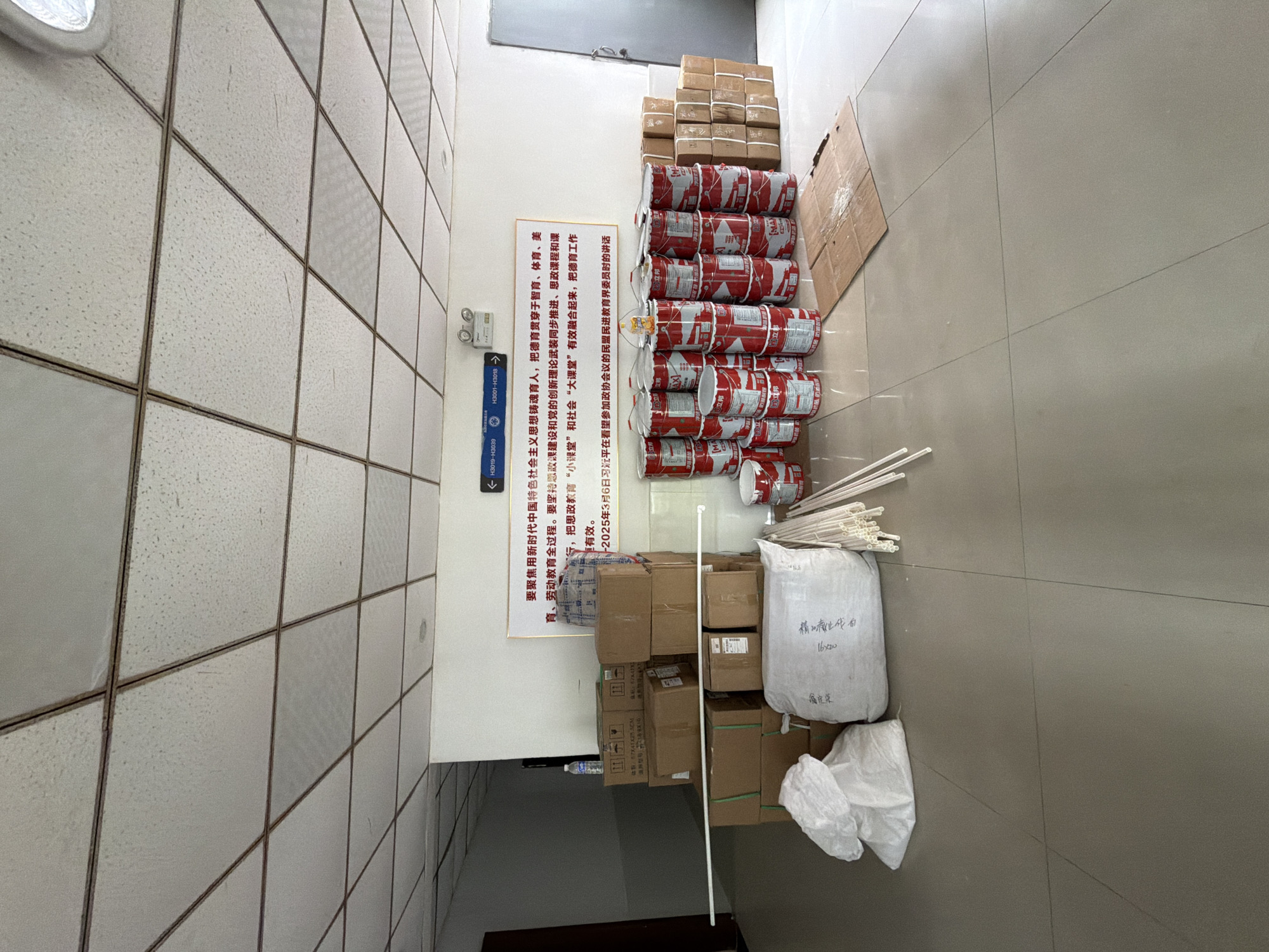 The image shows a hallway in what appears to be a school or administrative building in Jiangjin, China. In the center of the frame, a stack of cardboard boxes is piled on the floor, with some white plastic tubes and bags of material in front of them. To the right of the boxes, a row of red and white paint cans is arranged. A large white sign with Chinese text is mounted on the wall behind the boxes. The ceiling is a grid of acoustic tiles. The floor is tiled in a light beige color. There are no people visible in the image. The overall impression is one of an area where materials have been recently delivered or are being stored.