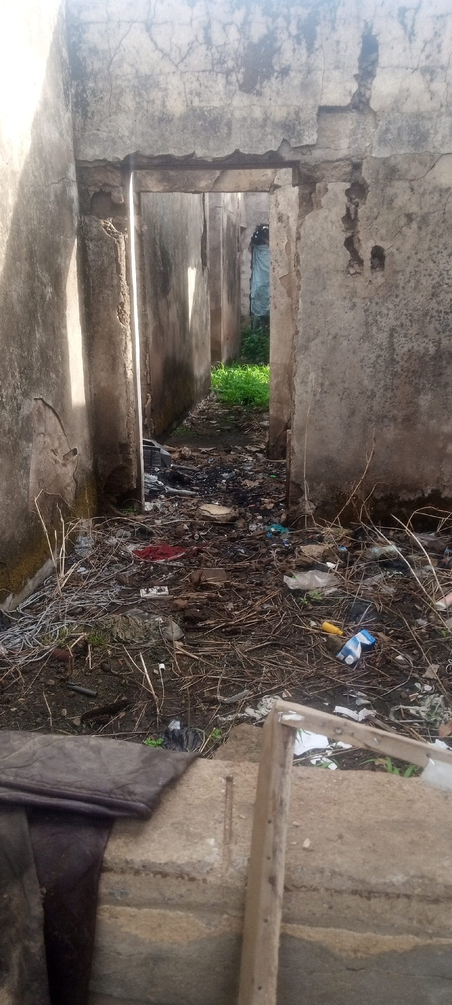 The image depicts an interior space of a derelict structure in Sabon Tasha, Nigeria. The building is constructed of concrete and displays significant deterioration, with crumbling walls and exposed sections. Multiple doorways or archways line a corridor filled with refuse, including what appears to be burned materials, dry brush, plastic debris, and other unidentified items. Vegetation is visible at the end of the corridor. In the foreground, there is what seems to be a concrete structure partially covered by dark material, alongside a wooden frame. The lighting is uneven, suggesting natural light entering the structure, illuminating some areas while others remain in shadow. The scene is indicative of neglect and abandonment.