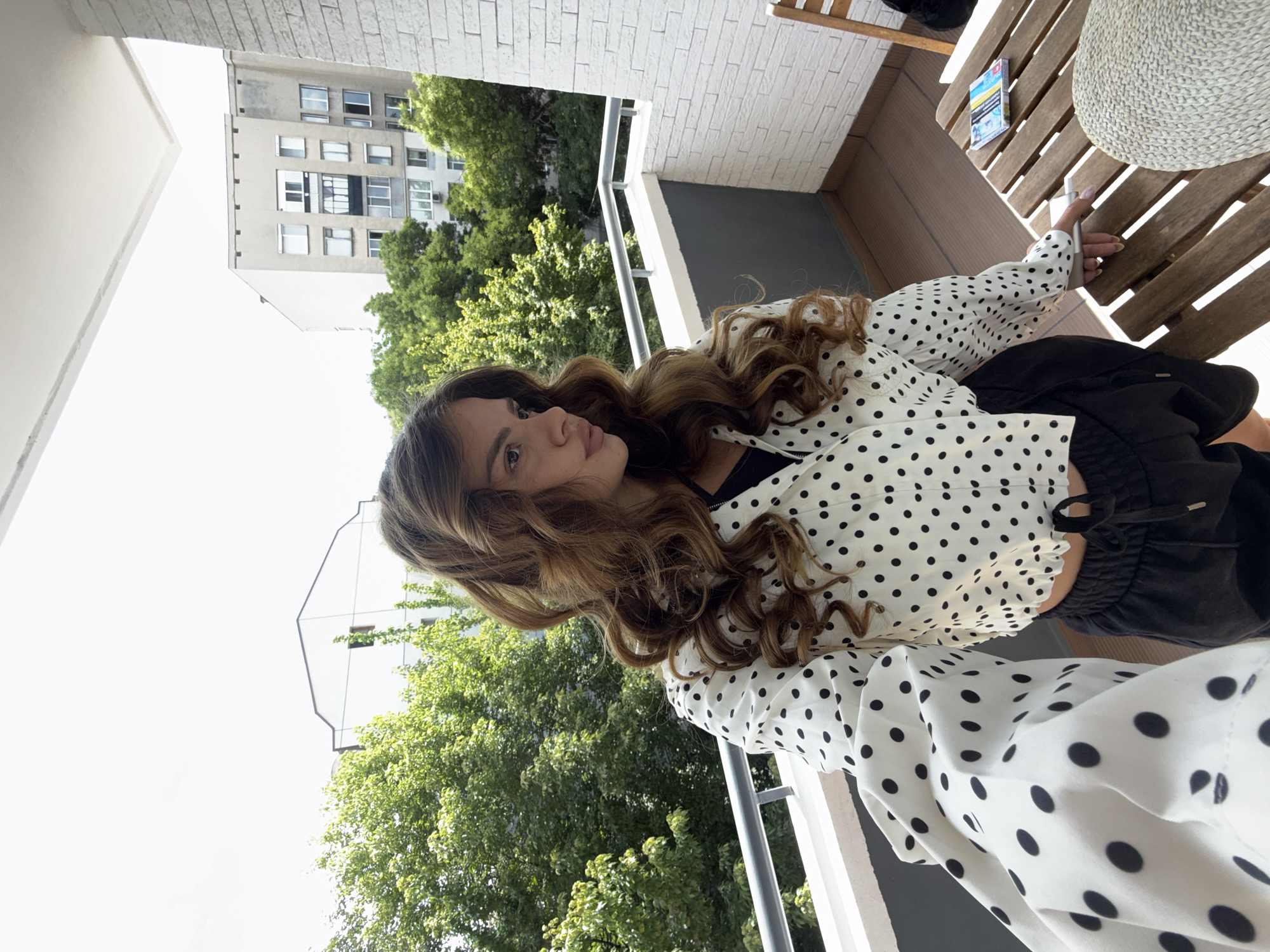A young woman stands on a balcony overlooking a residential area with trees and buildings. She has long, wavy brown hair and is wearing a white cropped jacket with black polka dots and black athletic shorts. She is looking to her left. On the wooden table to her right, there is a stack of books or a game, a pen, and a straw hat. The balcony railing is white, and the wall is made of white brick. In the background, there are green trees and apartment buildings. The sky is bright and overcast.