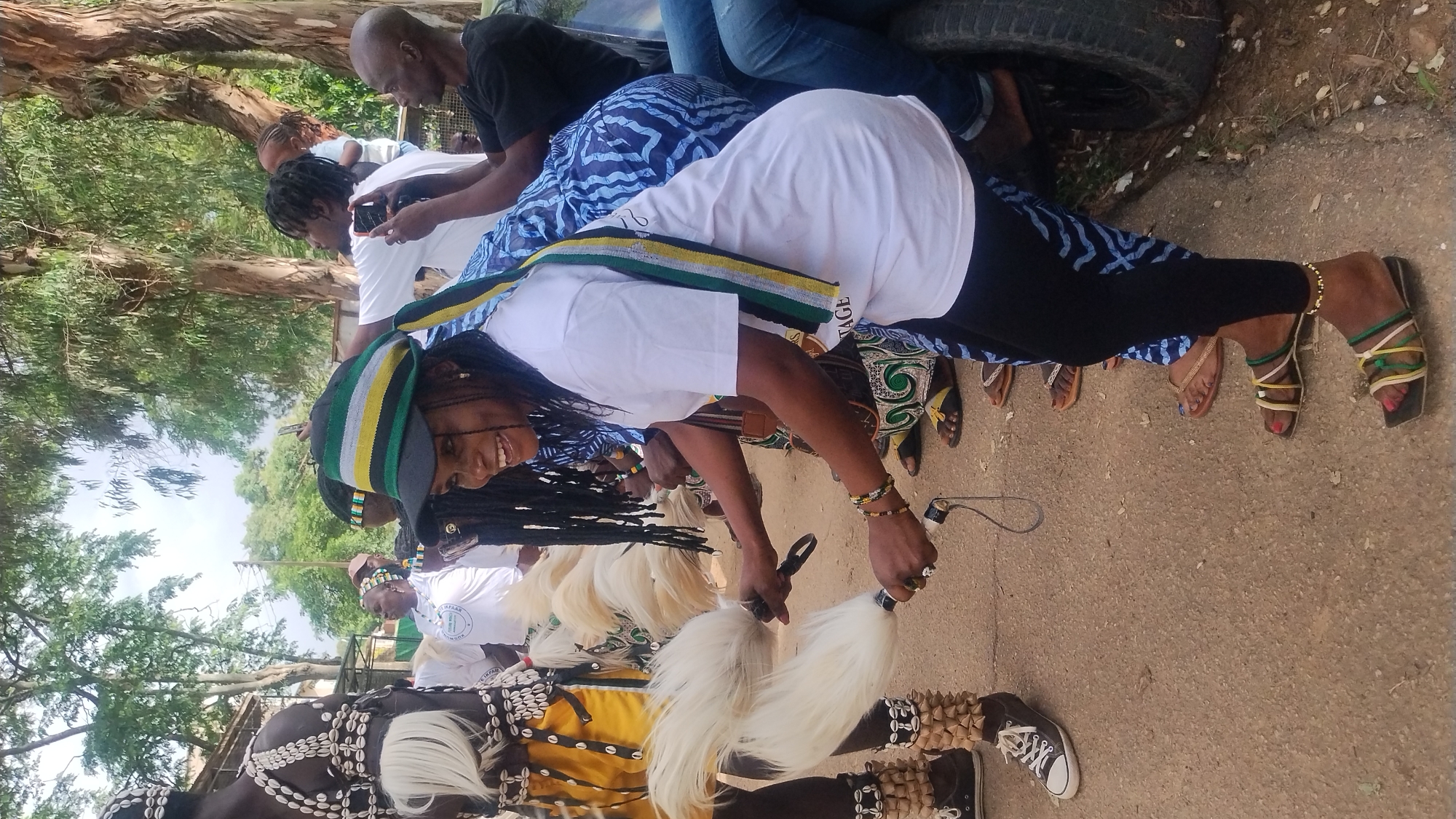 The image depicts an outdoor scene in Jos, Nigeria, where a group of people are gathered. In the foreground, a woman wearing a black and green striped hat and a white t-shirt is smiling and holding a white, feather-like object in each hand. She is wearing black leggings and multi-colored sandals. To her left, another person is partially visible, wearing a traditional outfit with yellow and white fabric, adorned with shells and white tassels.
Several other individuals are present in the background, some holding cell phones and appearing to take pictures or record the event. They are dressed in casual clothing, including t-shirts and patterned fabrics. The environment is dusty and appears to be a roadside with trees and vegetation. The overall atmosphere suggests a cultural event or celebration.